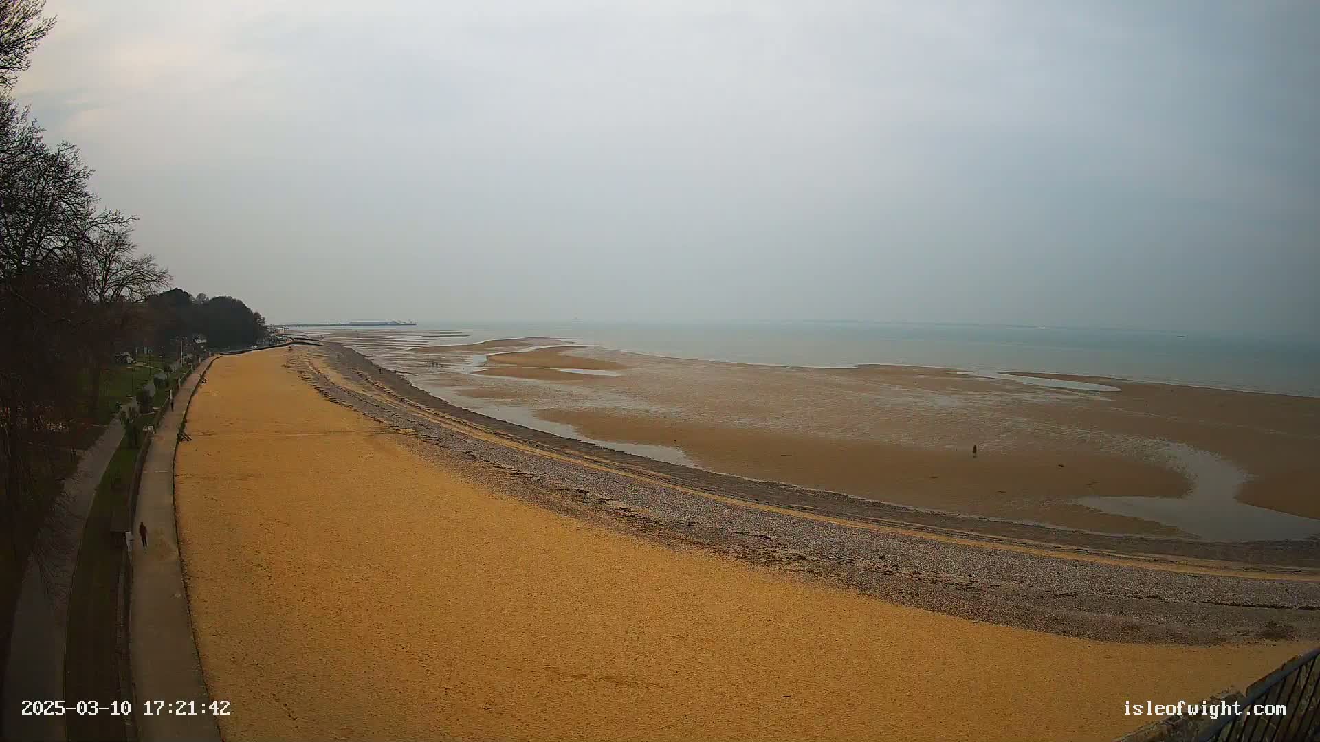 A sandy beach stretches along a coastline under a gray, overcast sky, with a paved walkway and trees visible in the background.