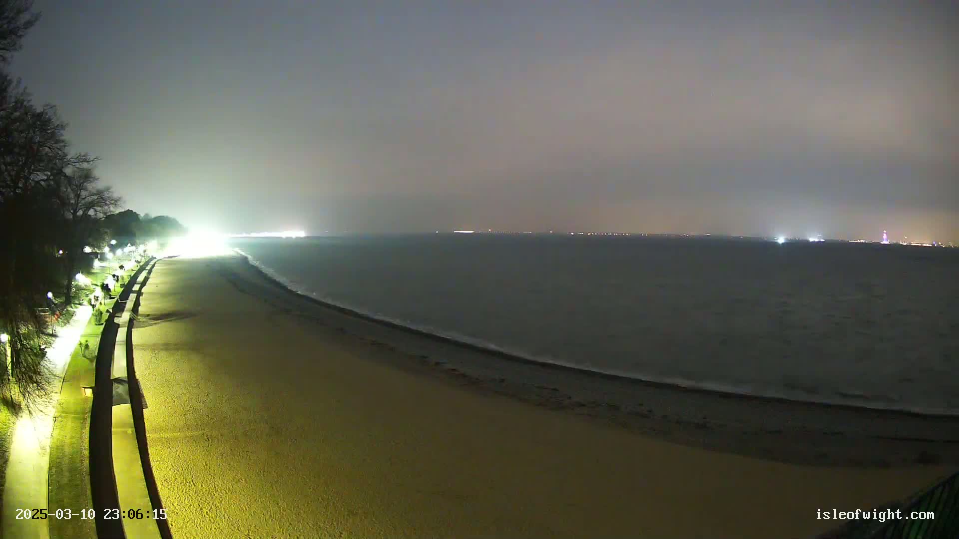 A nighttime view of a mostly empty sandy beach with a walkway and lights, overlooking a calm sea with distant city lights under a hazy sky.