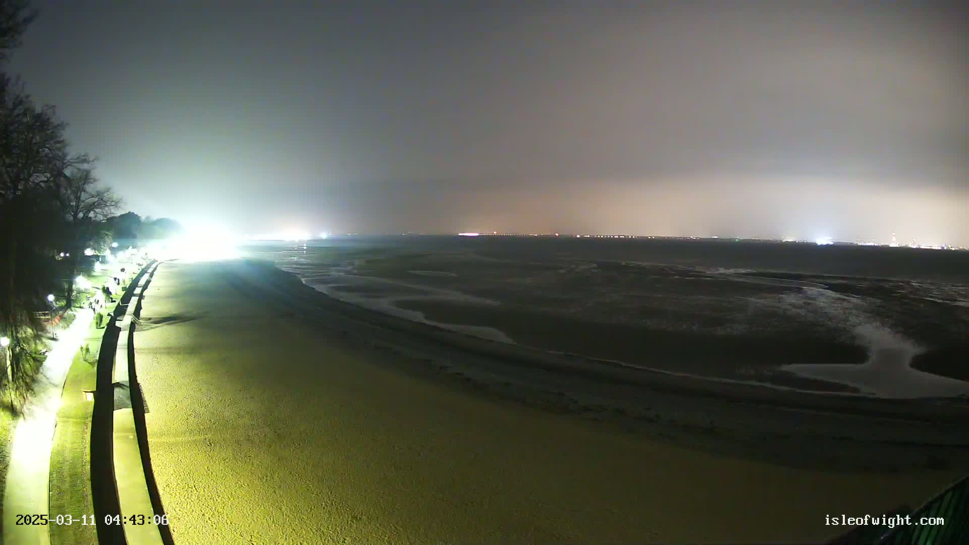 A nighttime view of a mostly empty beach and a paved walkway, illuminated by artificial lights, under an overcast sky.