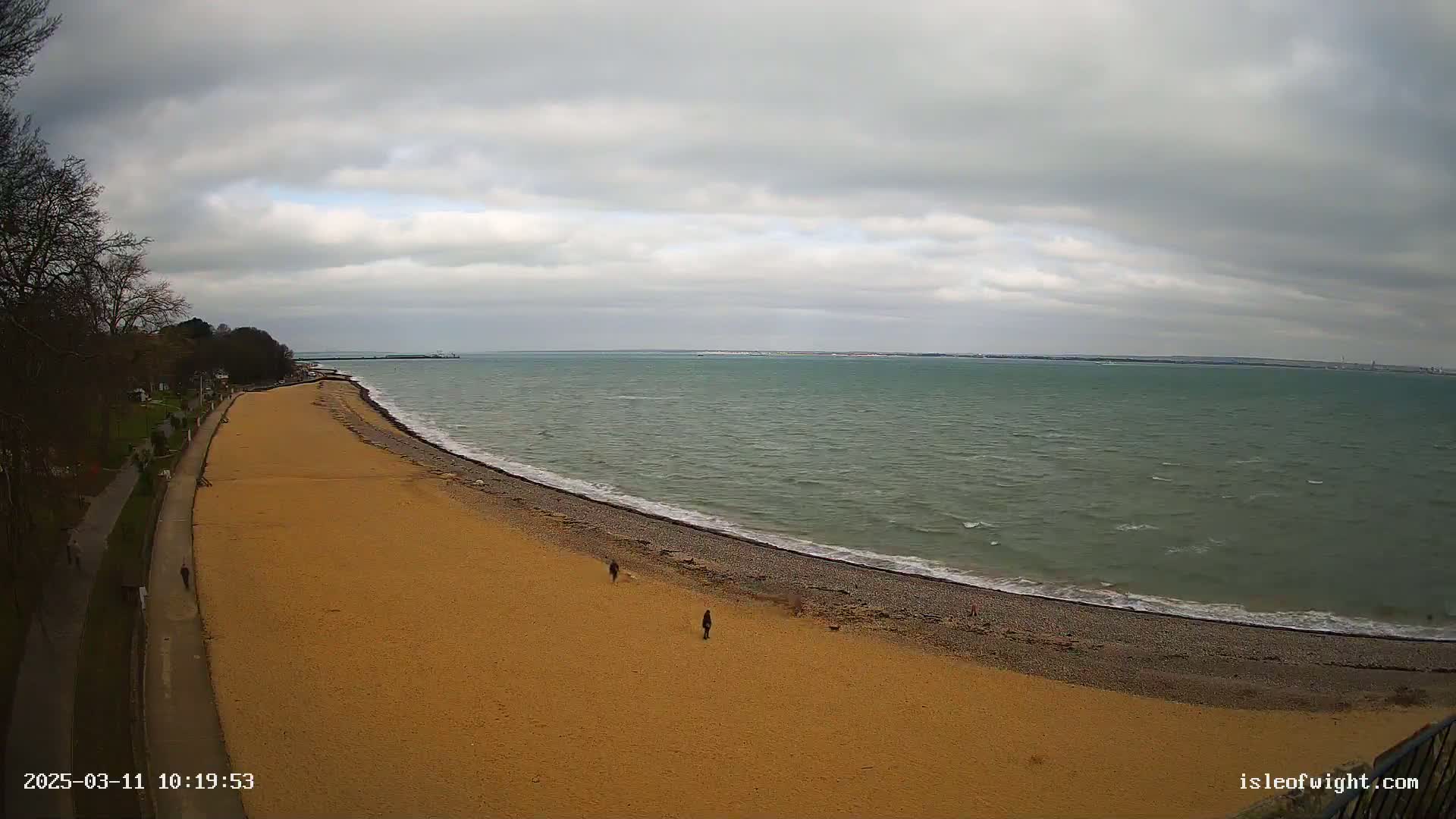 A wide, sandy beach curves gently into a calm, grey-green sea under a mostly cloudy sky.