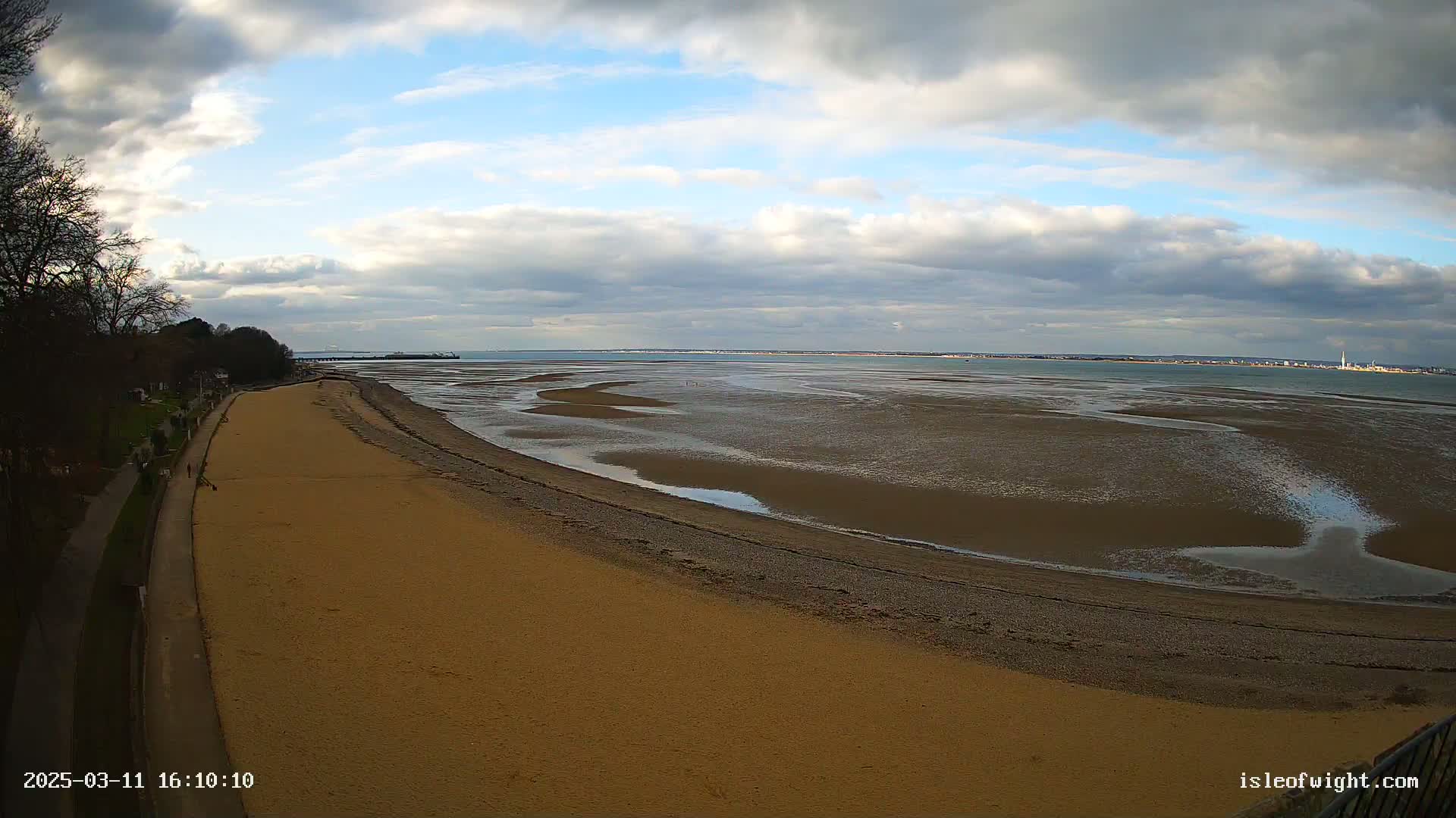 A sandy beach stretches alongside a paved walkway, leading to a wide expanse of exposed mudflats and calm water under a partly cloudy sky.