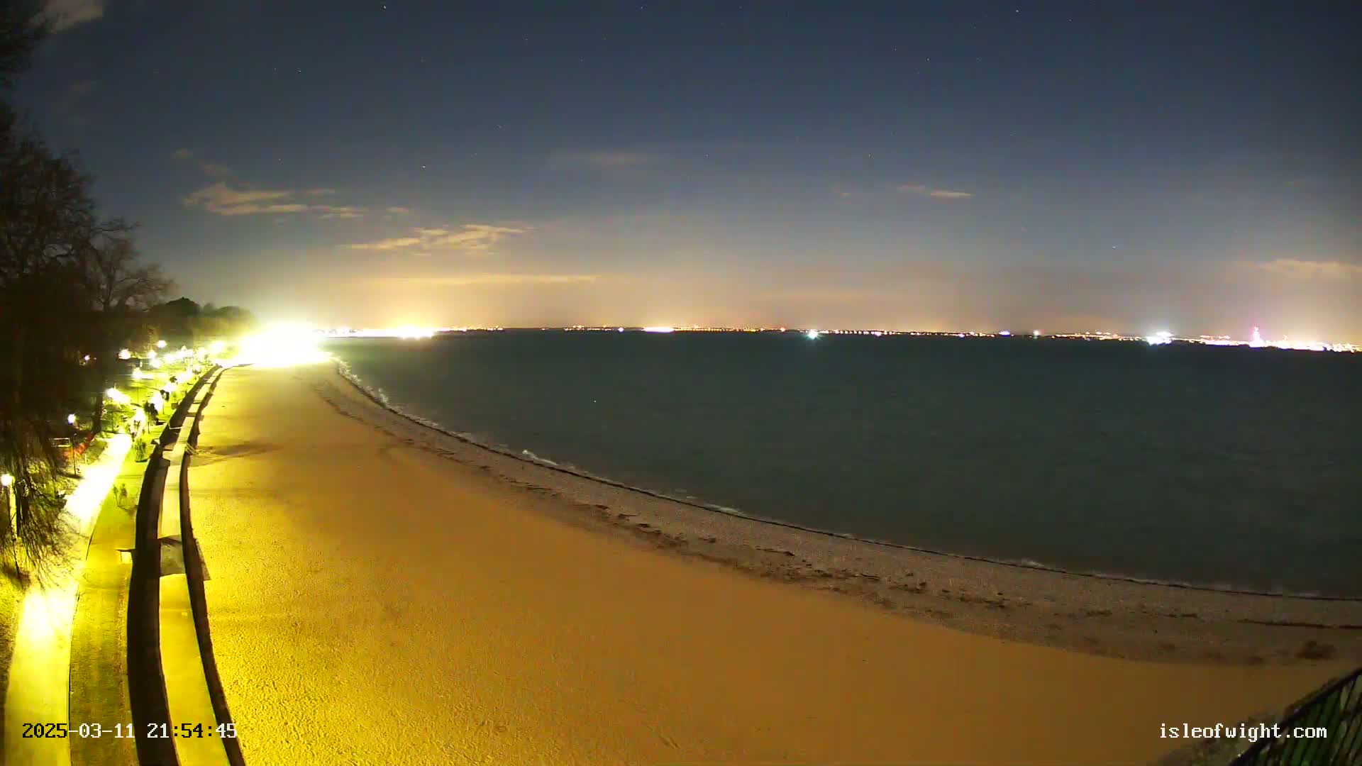 A nighttime view shows a sandy beach with a paved walkway lined with lights, gently sloping to a calm, dark-colored sea under a mostly clear, dark sky.
