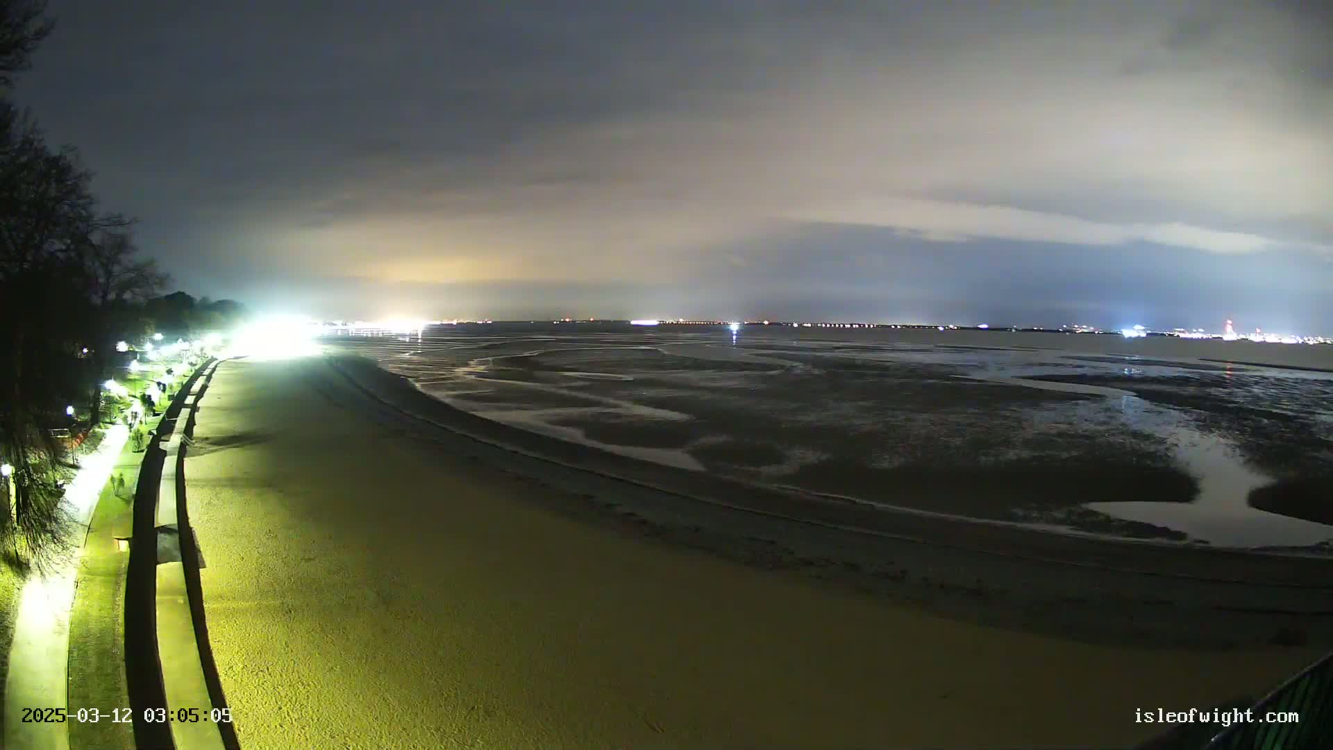 A mostly cloudy night scene shows a sandy beach at low tide, illuminated by lights along a seaside promenade, with a distant city skyline visible across the water.