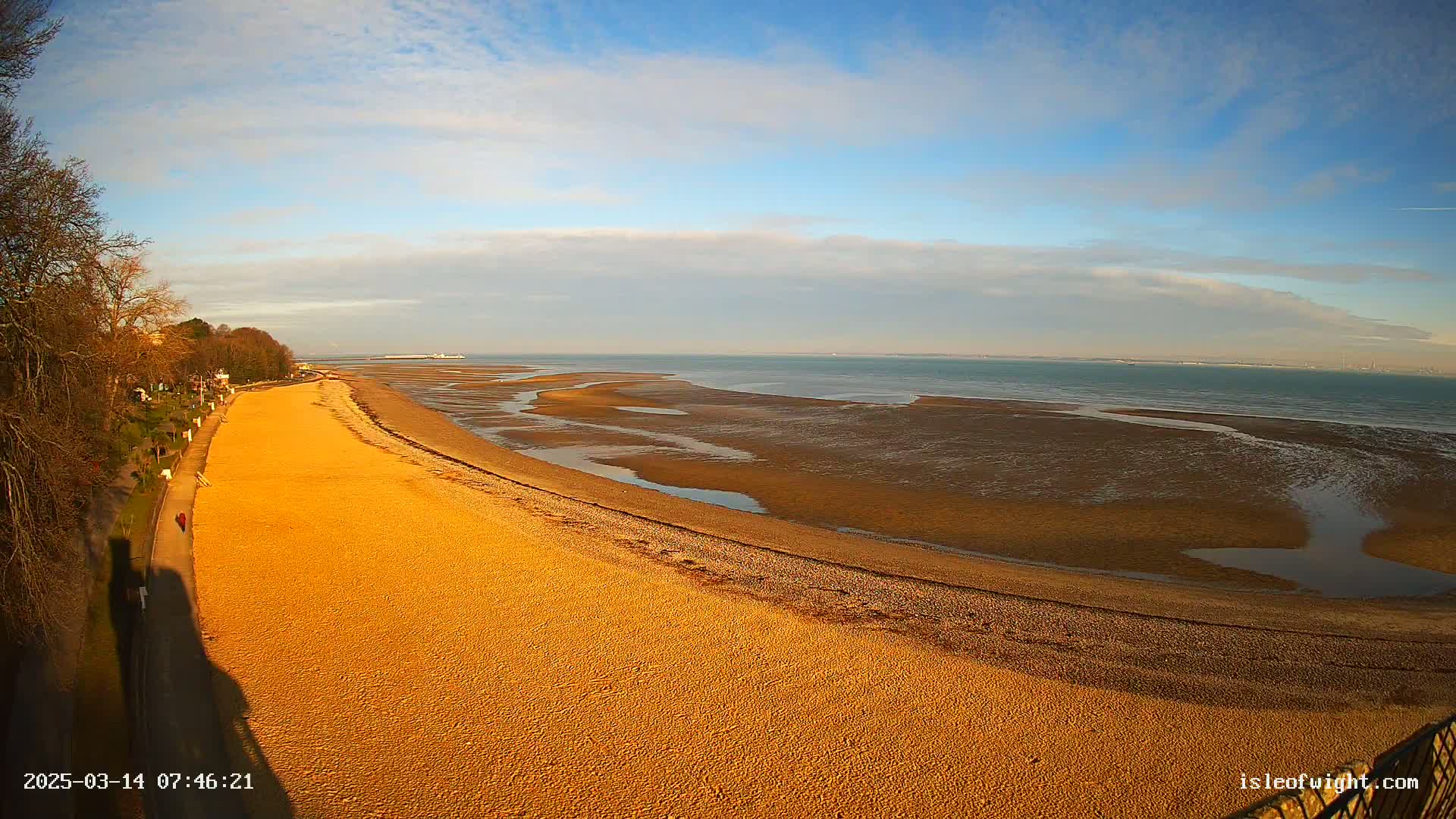 A long, sandy beach curves gently toward a calm sea under a mostly clear, sunny sky.