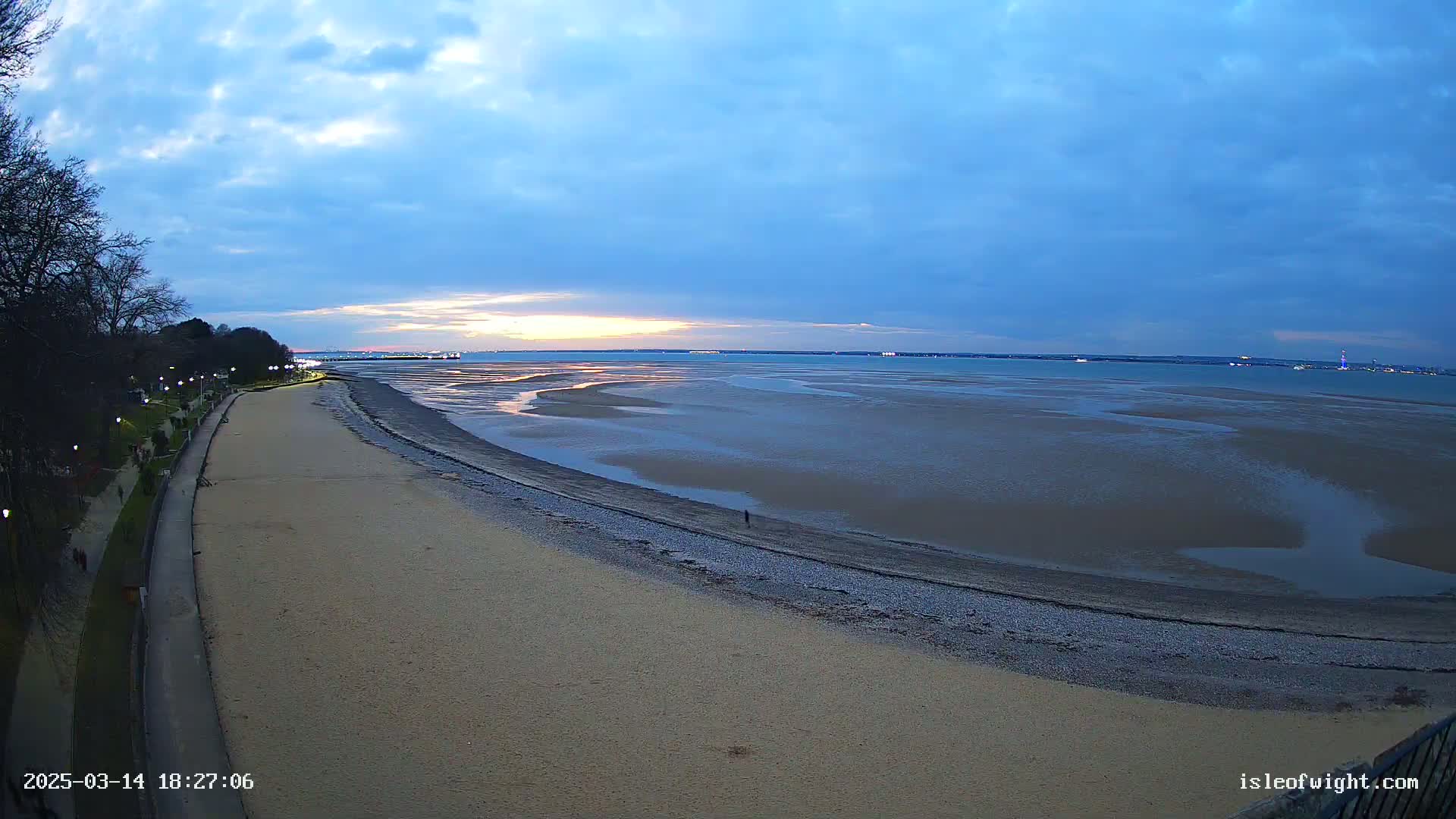 A sandy beach and paved walkway are next to a large expanse of exposed mudflats at low tide under a mostly clear, twilight sky.