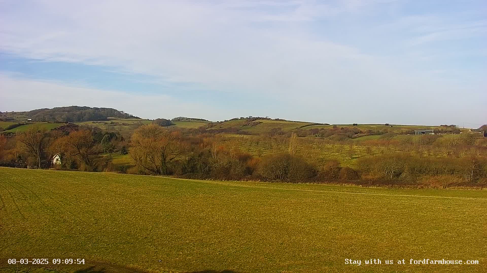A sprawling, grassy field stretches towards a line of low, rolling hills under a mostly clear, light blue sky.
