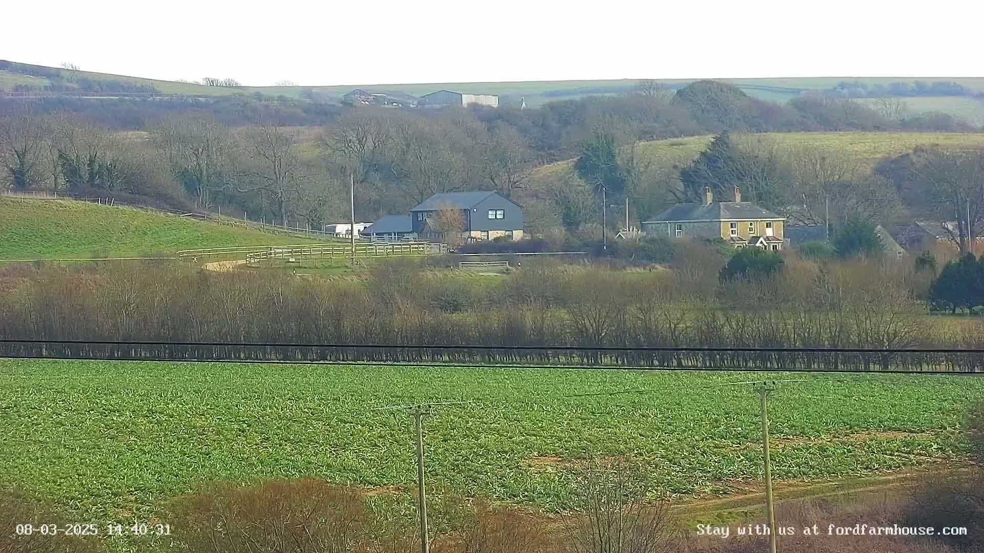 A sprawling green field sits in the foreground, beyond which are several buildings nestled among bare winter trees on rolling hills.
