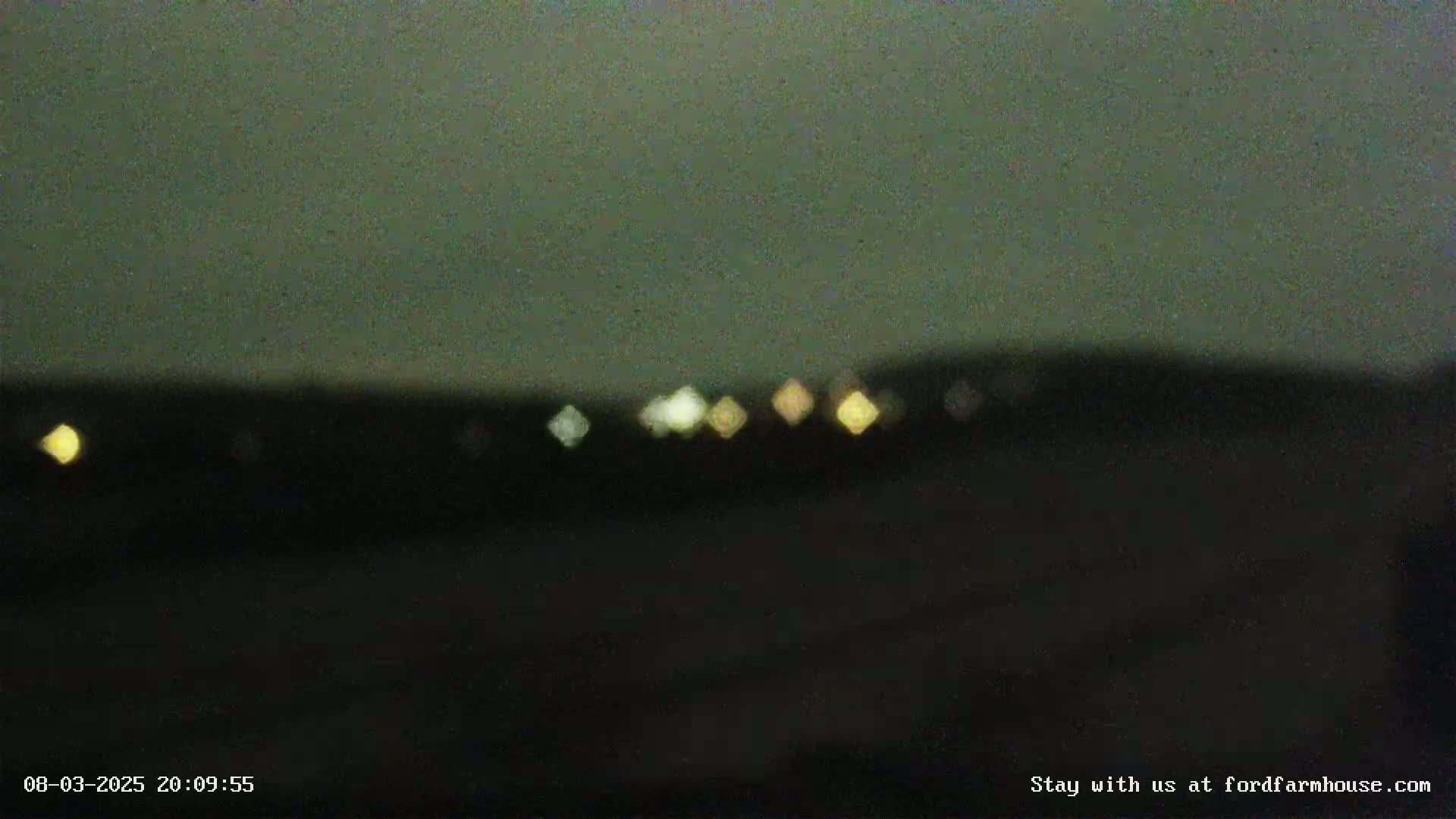 A blurry, nighttime image shows a dark landscape with several out-of-focus lights in the distance under a dark sky.