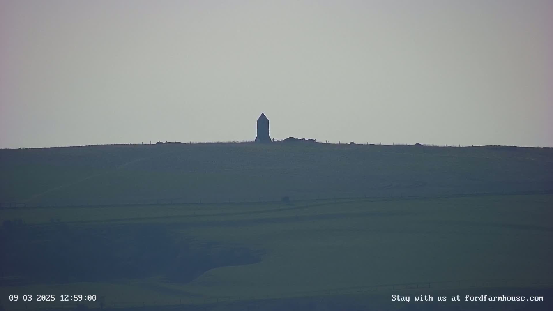 A dark, tower-like structure sits atop a gently sloping, green hill under a pale, overcast sky.