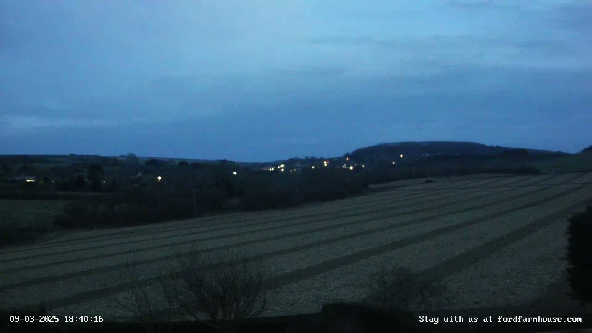 Under a dark blue twilight sky, a plowed field stretches towards a distant line of lights nestled in a low hill.