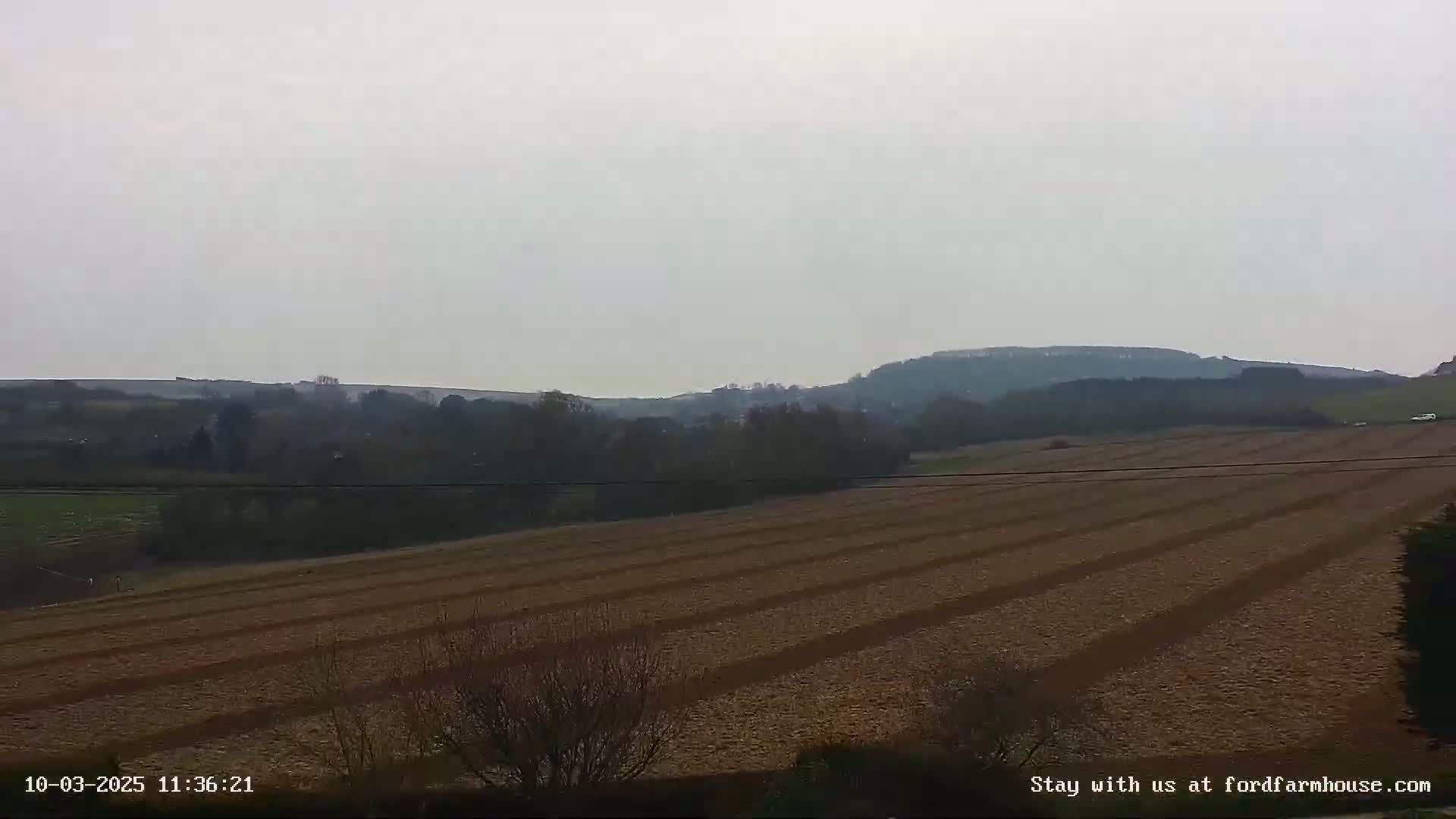 A plowed field stretches to a line of trees and distant hills under a hazy sky.