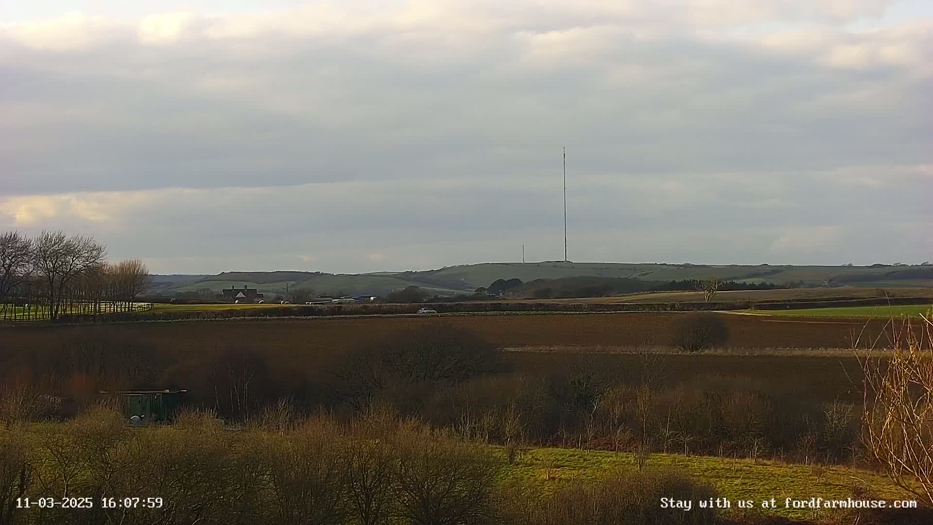 A flat, brown farmland landscape stretches out under a cloudy sky, with a tall antenna visible on a distant hill.