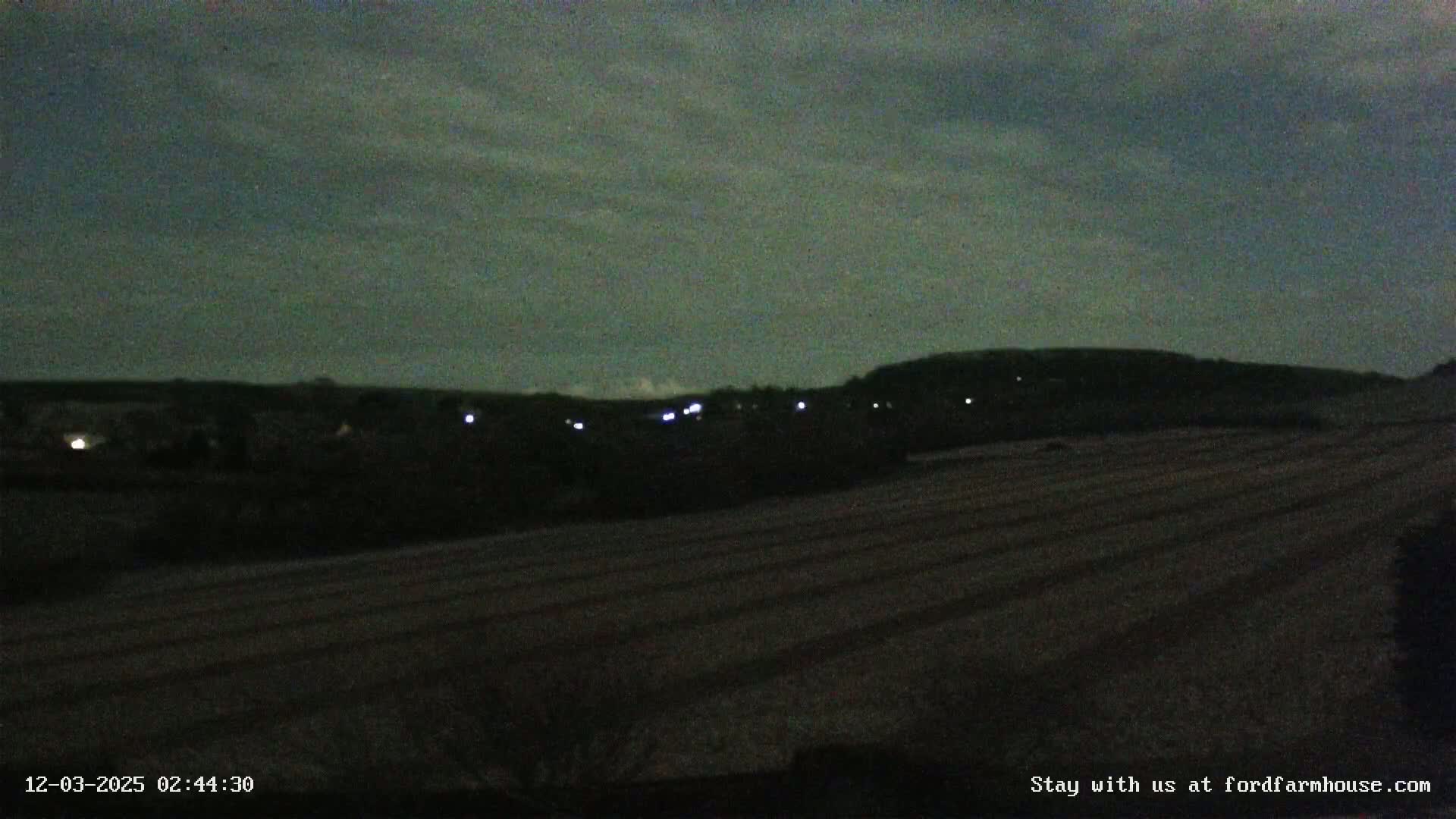 A dark, nighttime landscape shows a field in the foreground, a line of dimly lit buildings in the midground, and a hill in the background under a mostly cloudy sky.