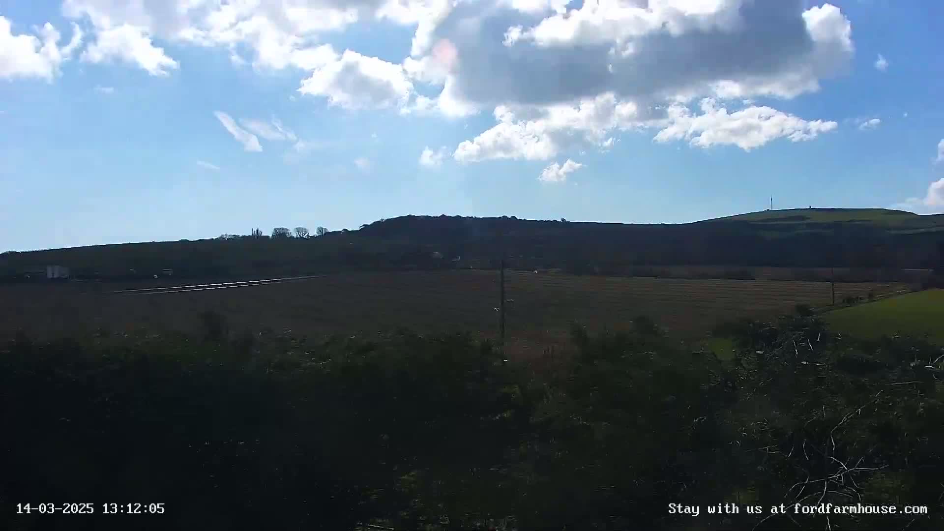 A partly sunny day reveals a view of rolling hills and fields, with dark shrubbery in the foreground.