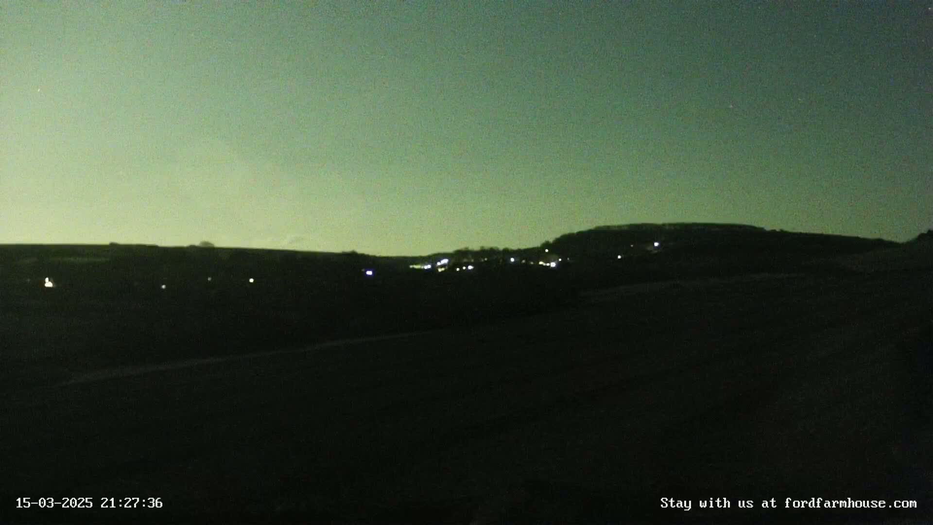 A nighttime, low-light view of a dark landscape with a hill in the distance showing faint lights, under a dusky greenish sky.