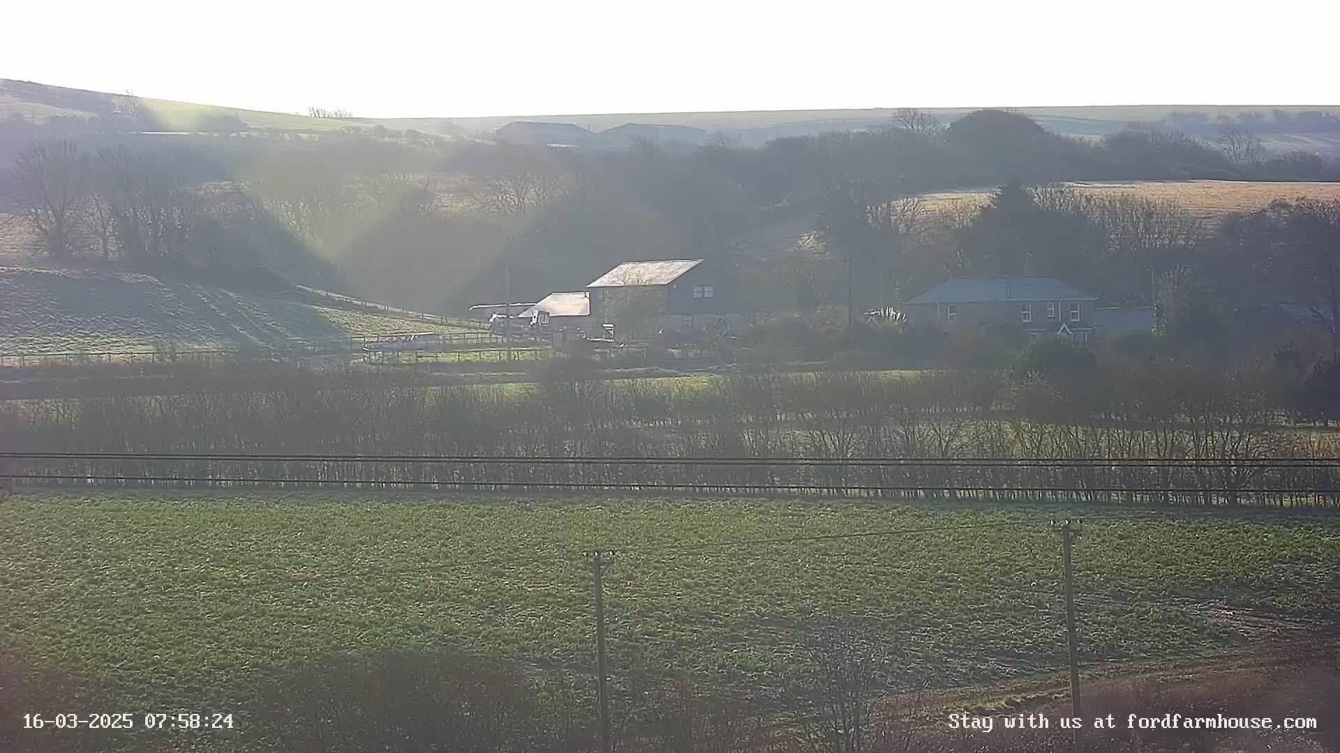 A hazy sunlit rural landscape features two buildings, one a large barn and the other a smaller house, nestled amongst trees and fields.