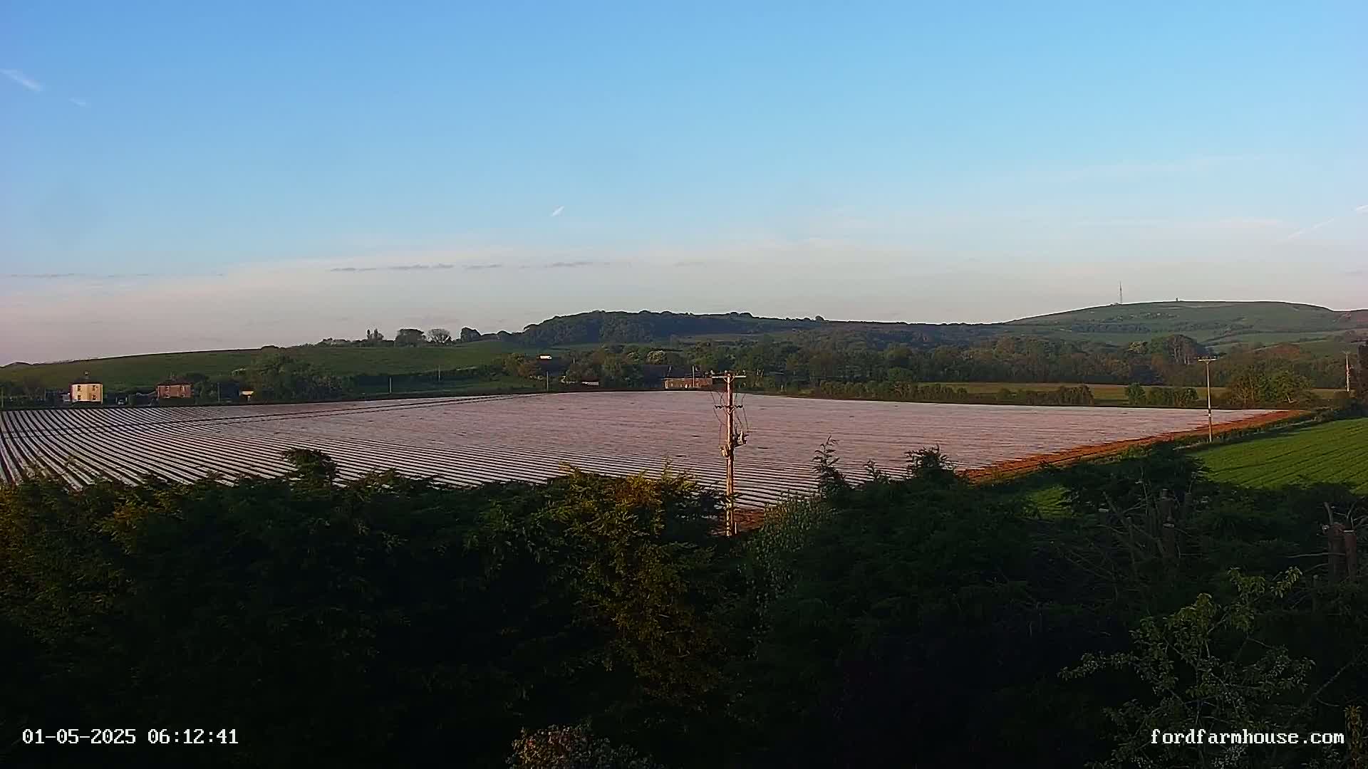 A large agricultural field, covered in plastic sheeting, stretches across a gently rolling landscape under a clear, pale blue sky.