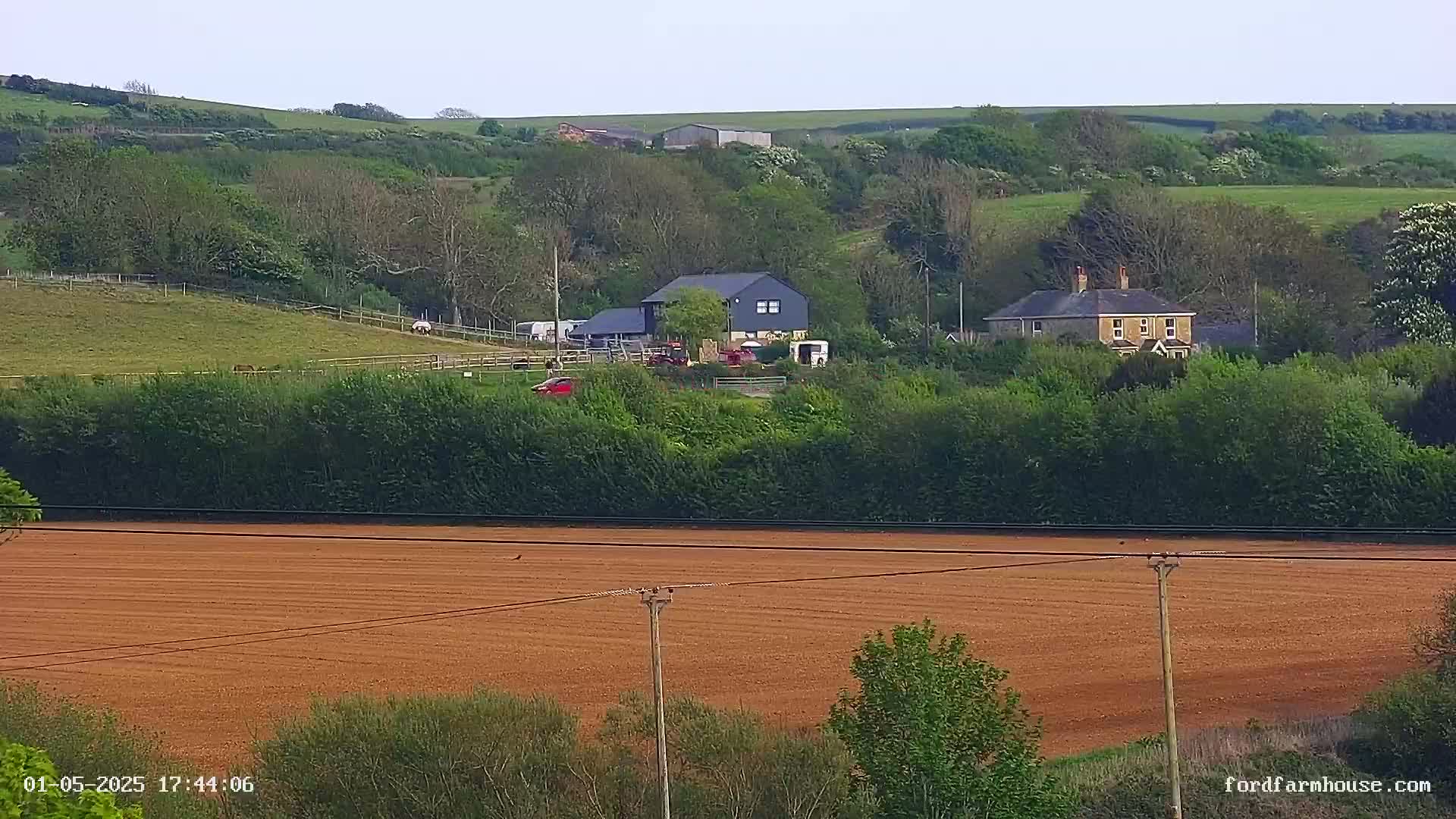 A sprawling farm scene, under a partly cloudy sky, features several buildings nestled among green hills and a large, tilled brown field in the foreground.