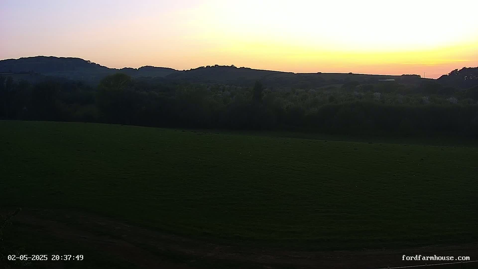 A sunset casts a warm glow over a dark green field, silhouetting a line of trees and distant hills.