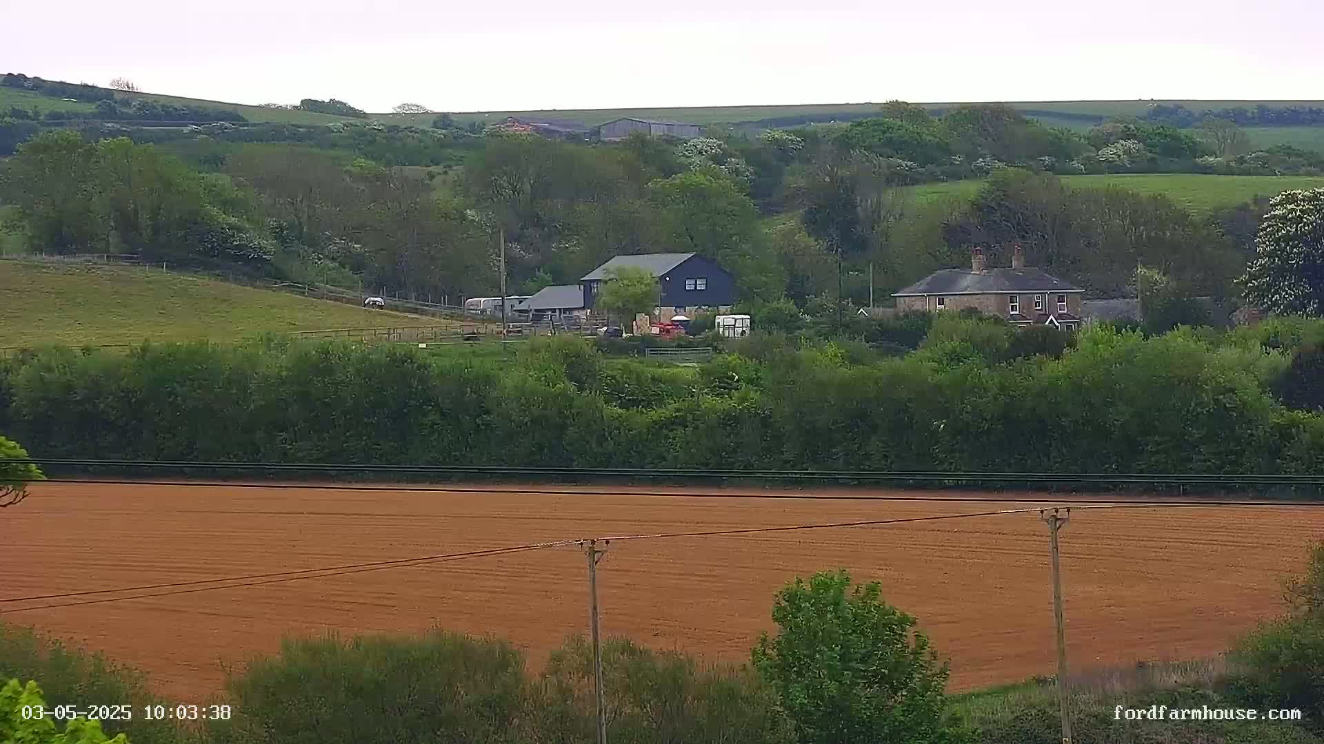 A sprawling farmland landscape under a cloudy sky features a dark-colored barn, a stone house, and a large tilled field in the foreground.