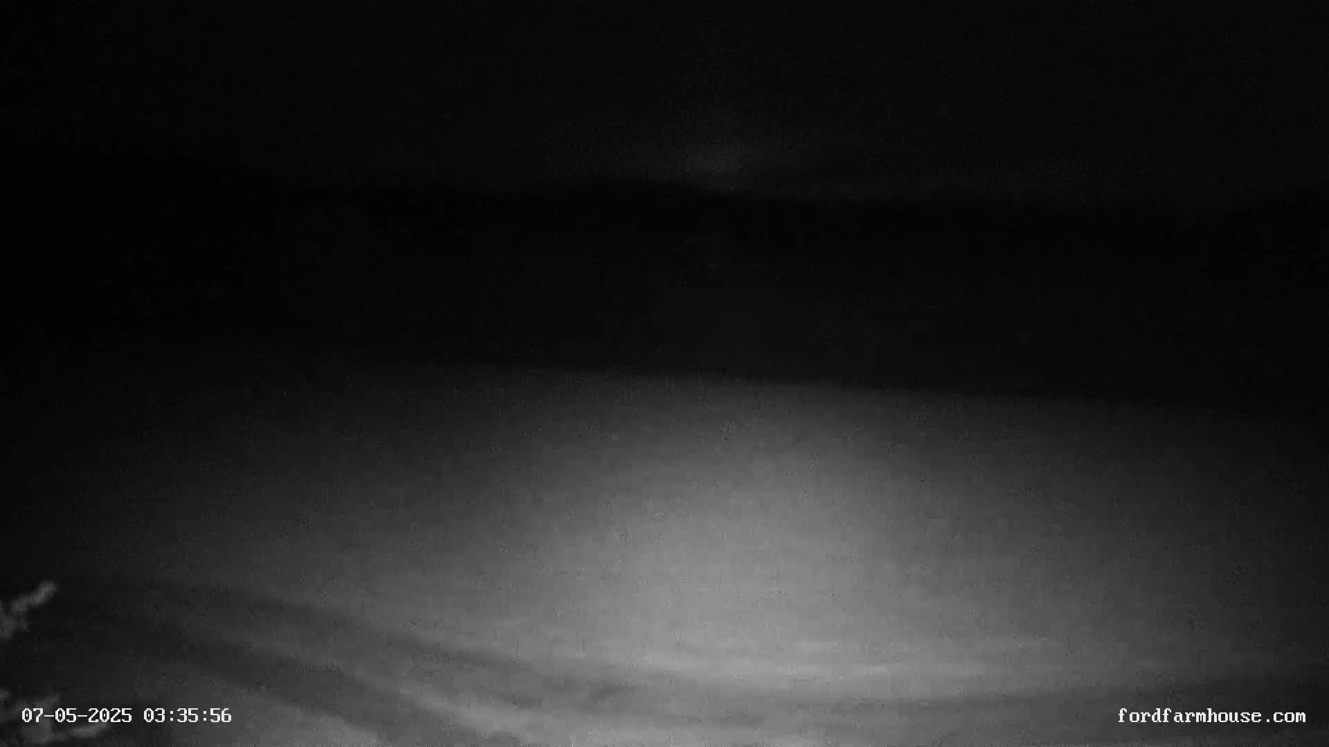 A low-light, nighttime outdoor image shows a mostly dark sky with some faint cloud cover and a dimly lit ground.