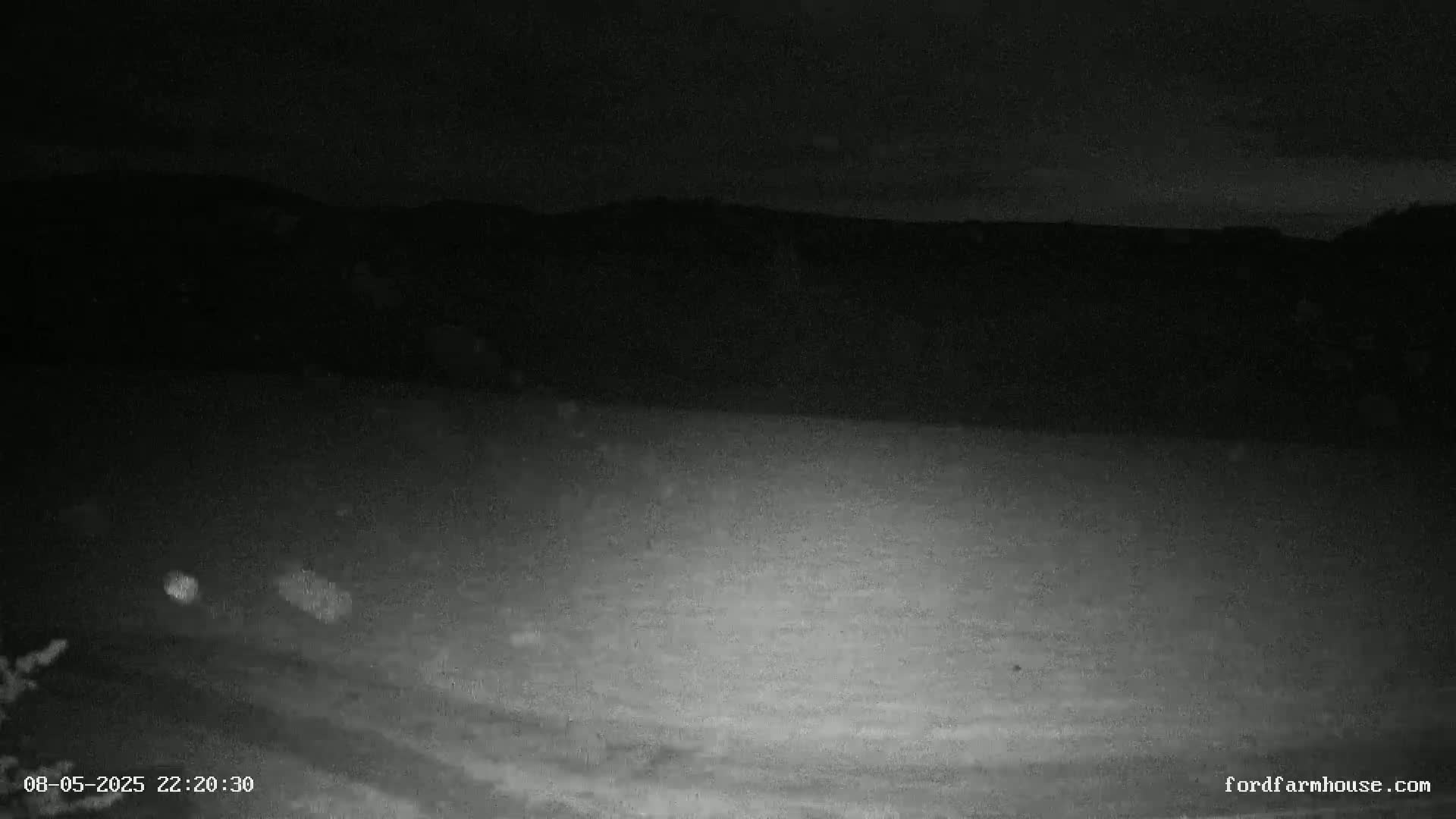A nighttime, low-light image shows a dark field with a dimly lit horizon and distant hills.