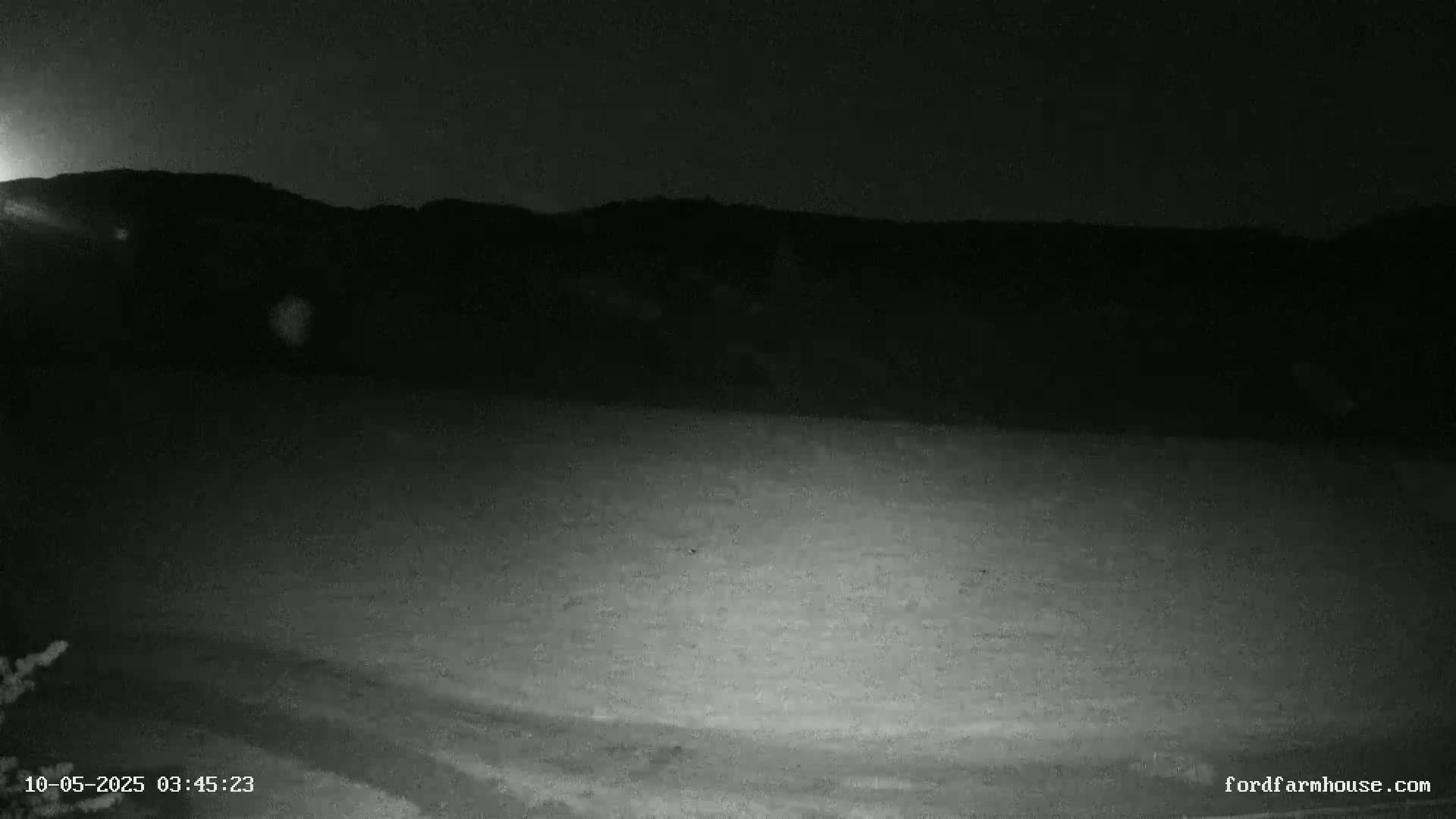 A nighttime, low-light view shows a flat, dark expanse of land extending to a dark, silhouetted mountain range under a clear, dark sky.
