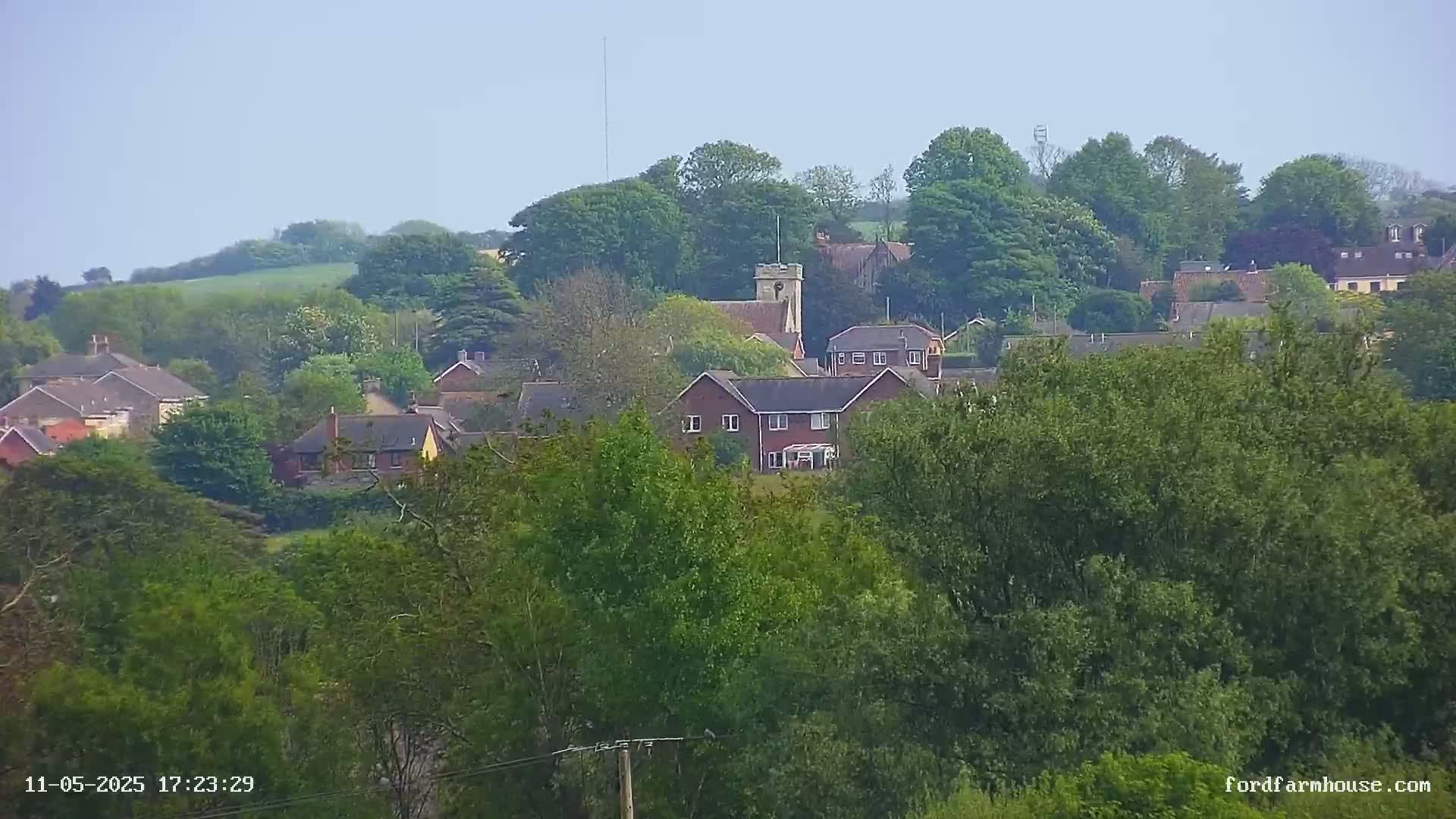A hazy view of a village nestled amongst lush green trees and rolling hills, under a clear sky.