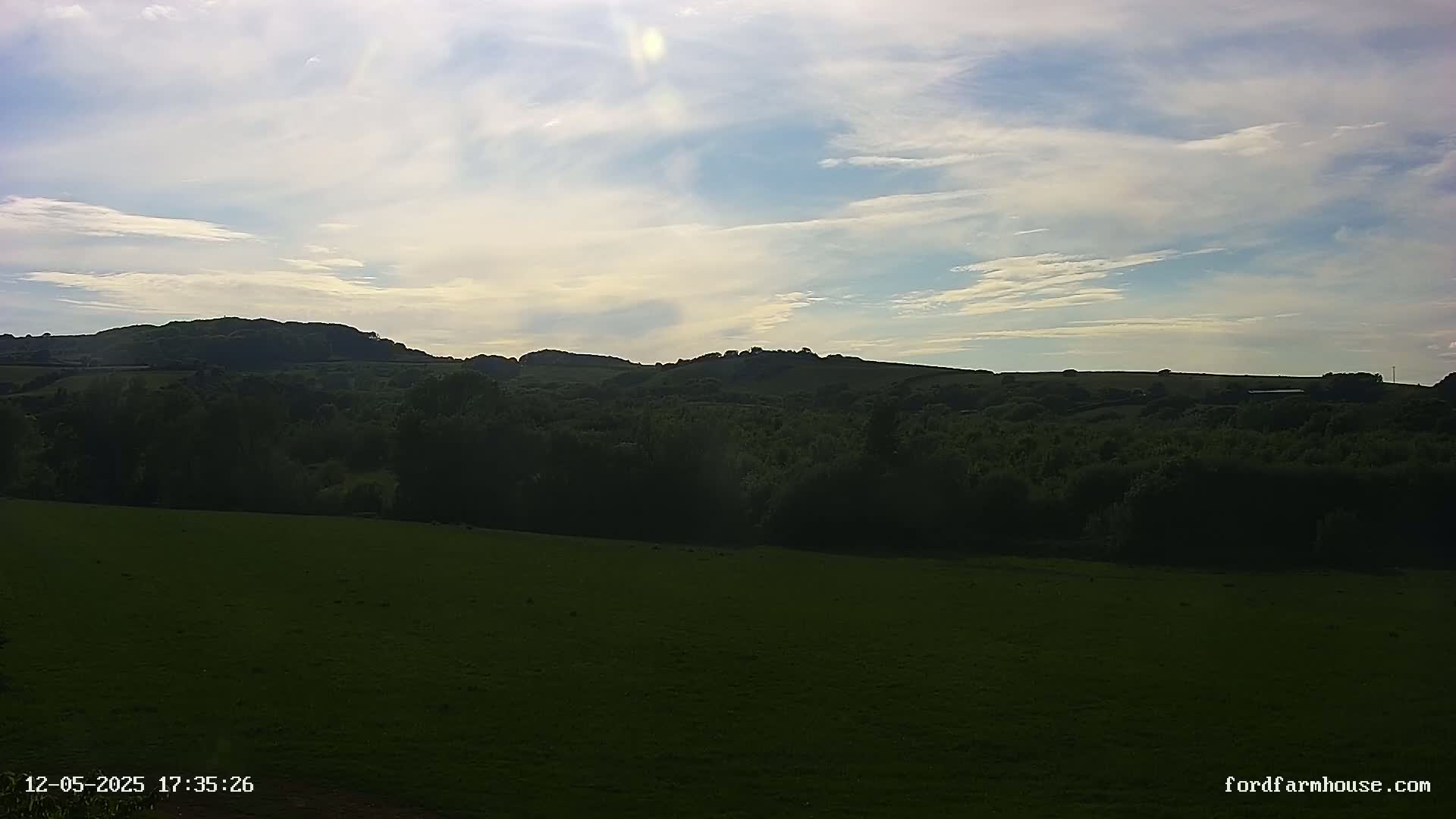A grassy field stretches out to a line of dark green hills under a partly cloudy sky.
