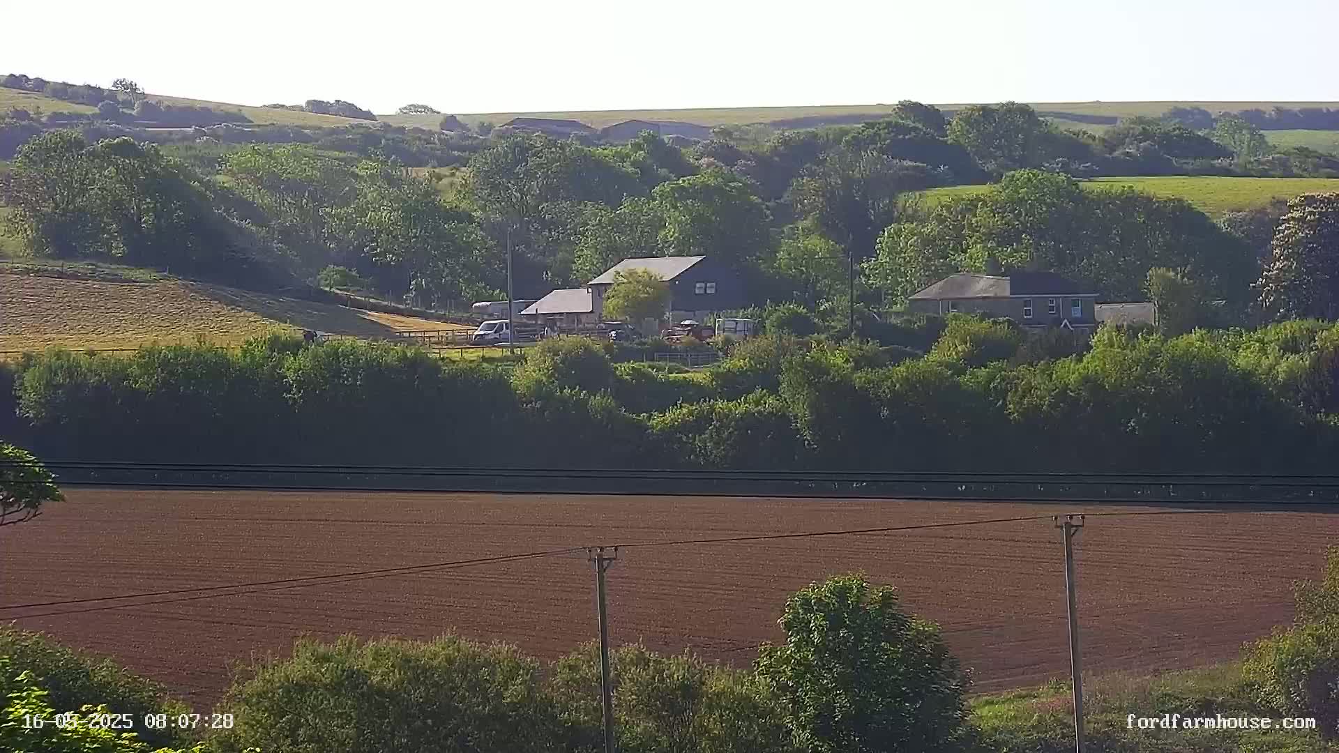 A large, dark-colored building and a smaller house sit nestled among lush green trees and hills on a sunny day, overlooking a wide, dark brown plowed field.