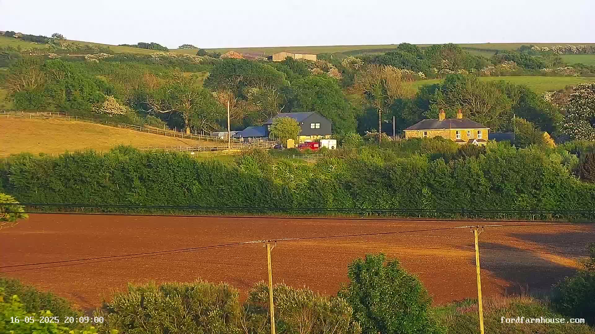 A sprawling farm scene with two houses, a dark-colored barn, and a reddish-brown field under a clear, bright sky.