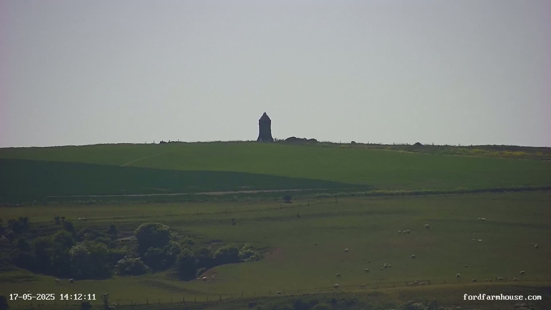 A dark, tall structure stands atop a green hill under a pale sky, with green fields and a few sheep visible in the foreground.