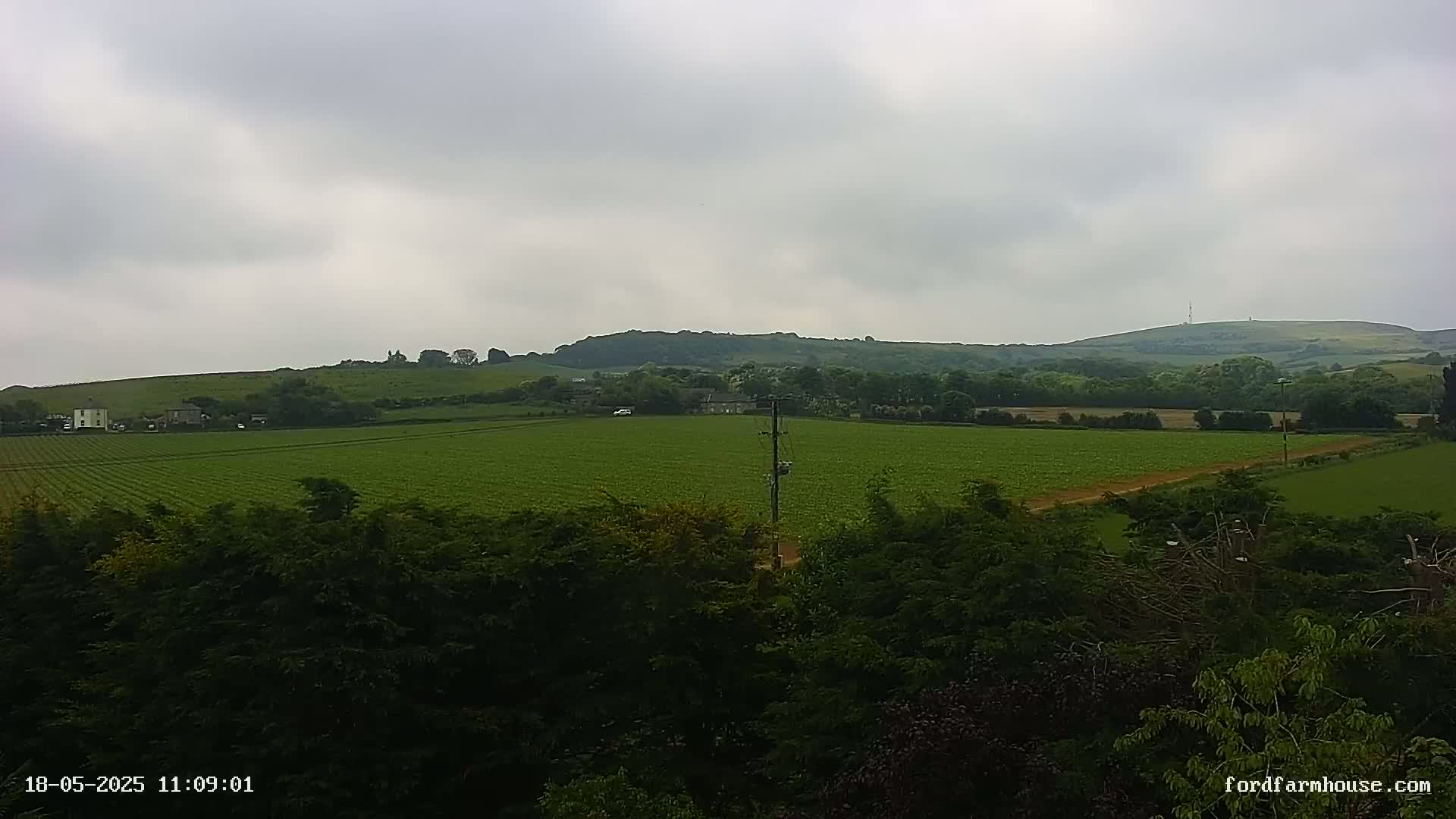A sprawling green field, dotted with a few buildings, extends to rolling hills under a cloudy sky.