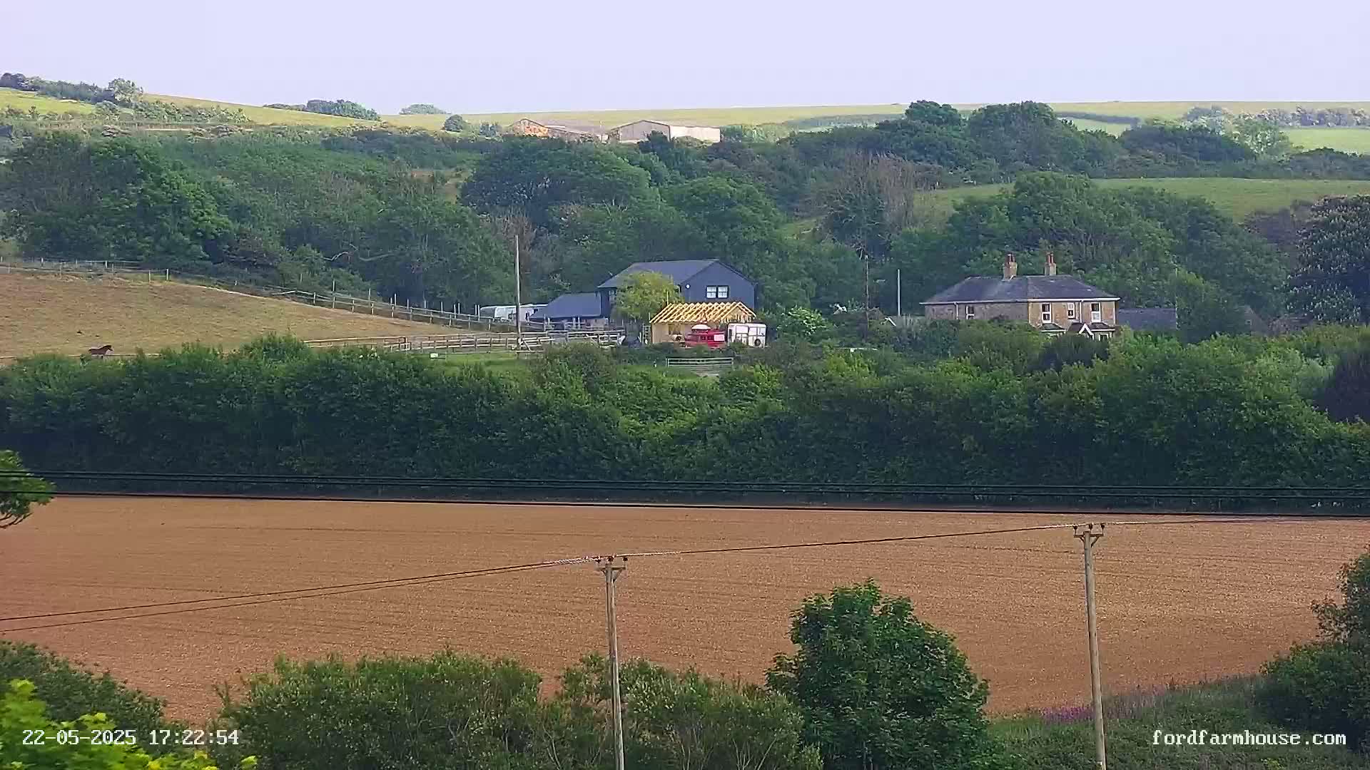 A brown field stretches in the foreground, beyond which sits a farmhouse and outbuildings nestled among green hills under a cloudy sky.