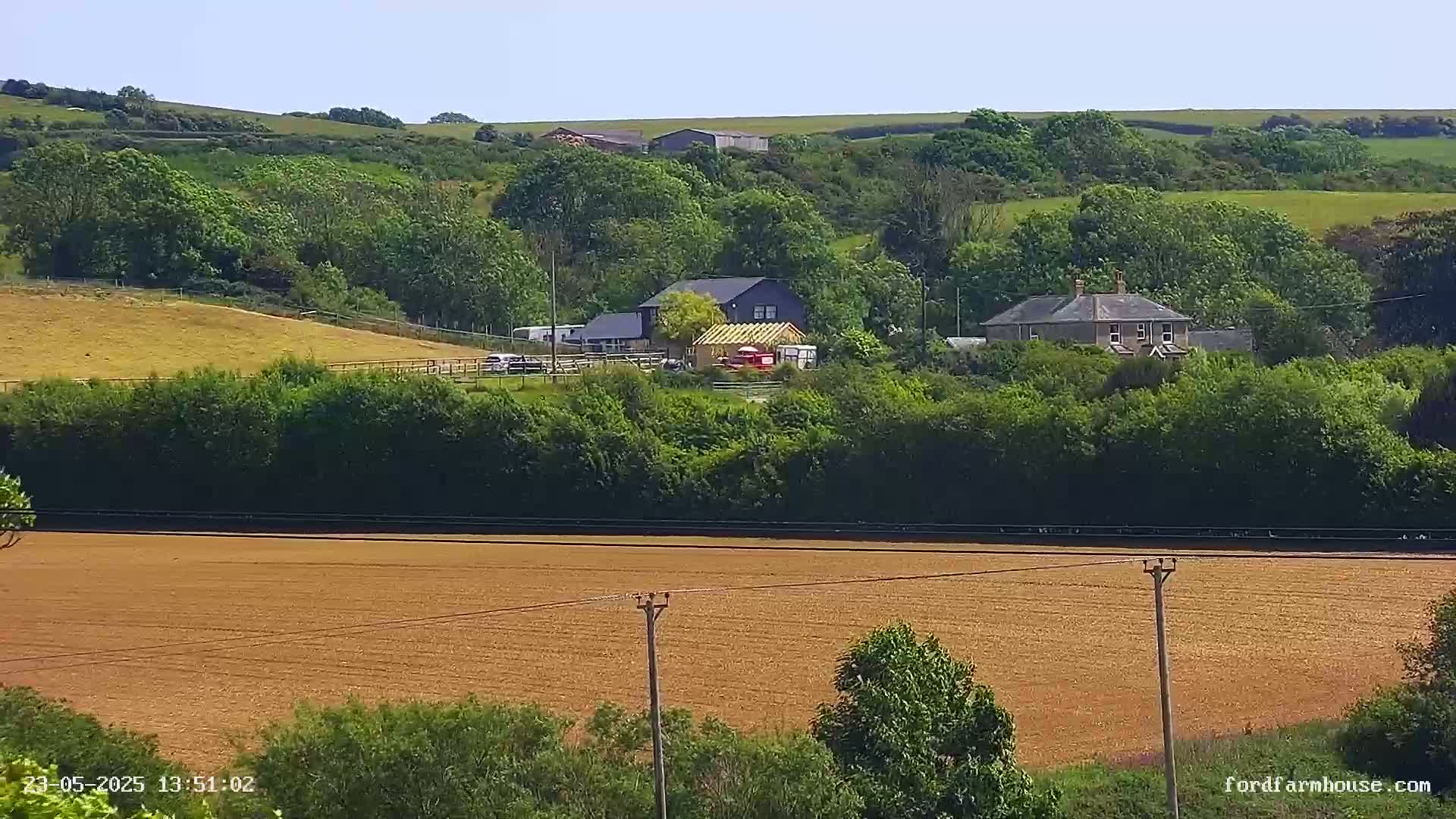 A long brown field stretches in the foreground, with a line of trees and two houses nestled among greenery on a hillside in the background under a clear sky.