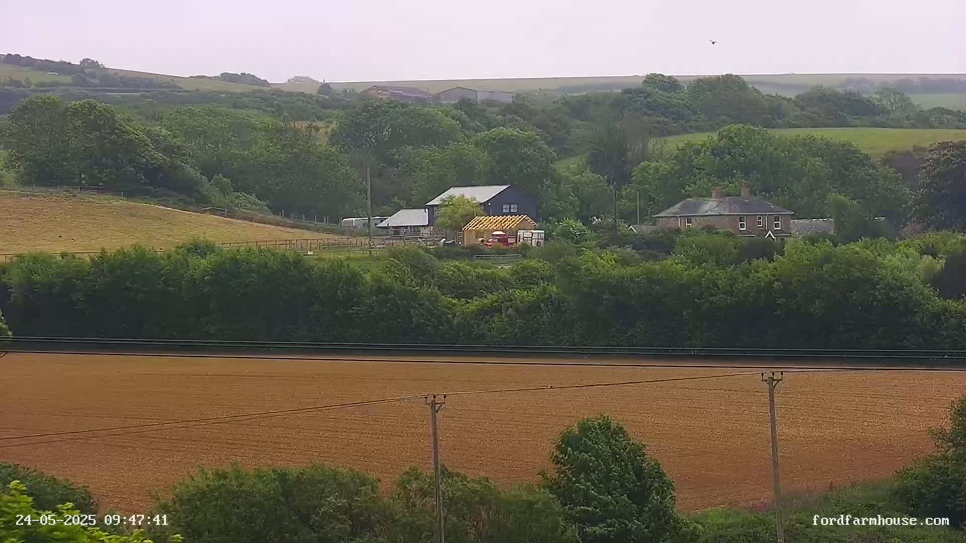A tan field sits in the foreground, with a line of trees separating it from a rural landscape featuring two houses and a building under construction, all under an overcast sky.