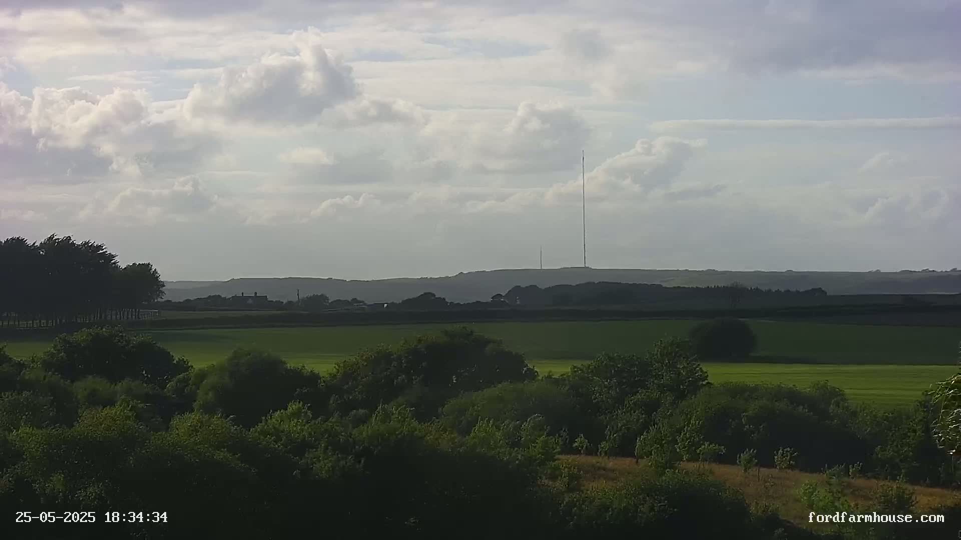 A partly cloudy sky overlooks a vast, green field dotted with trees, and a tall radio tower stands prominently on a distant hill.