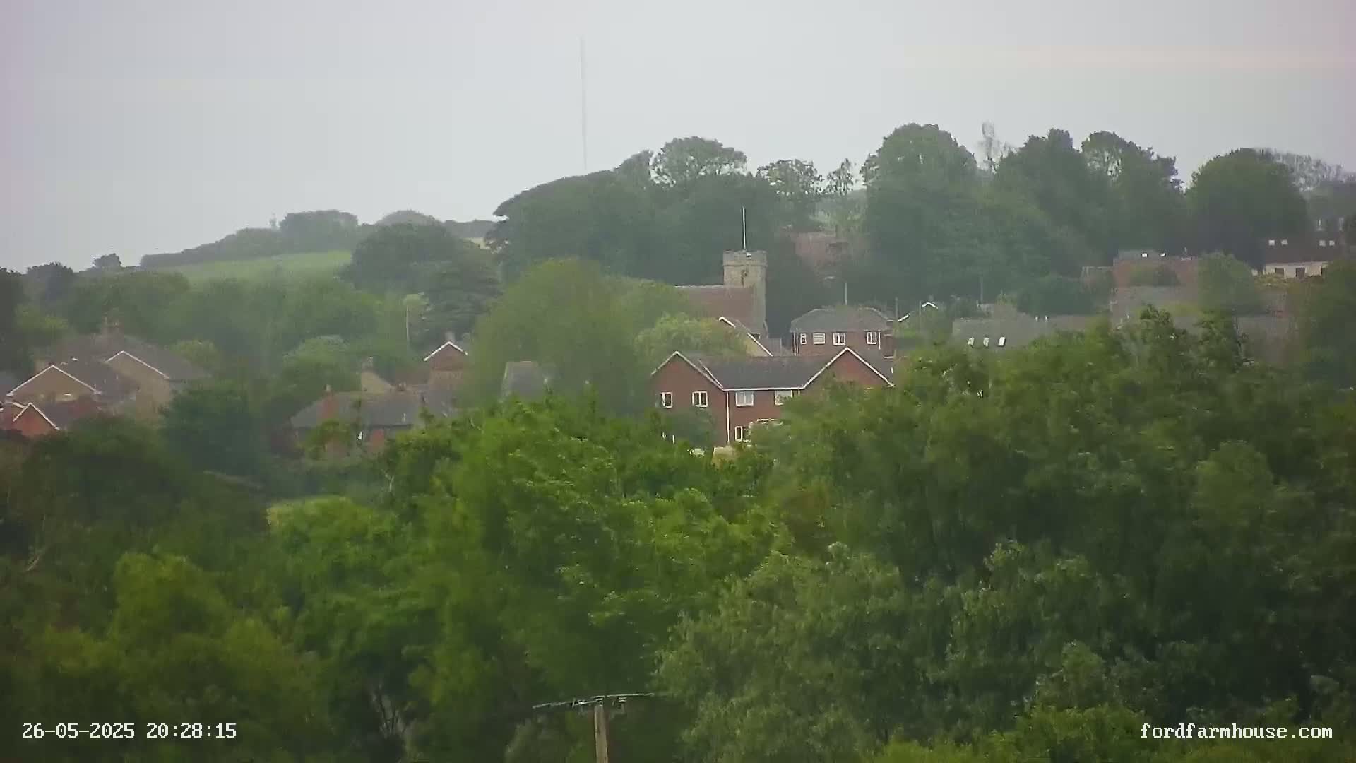 A small town nestled among lush green trees and hills under an overcast, rainy sky.