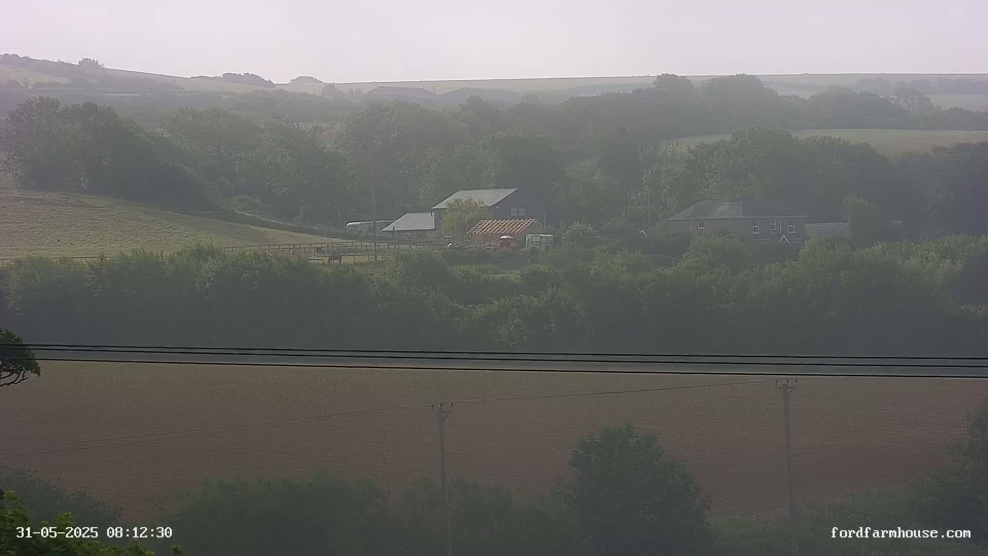 A hazy, distant view of a rural landscape shows farm buildings nestled among green hills and fields.