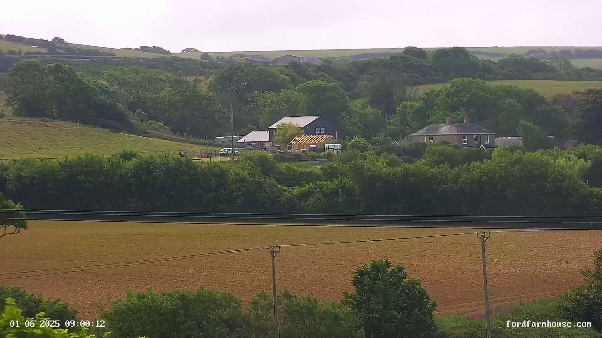 A rural scene under an overcast sky shows a farmhouse and outbuildings nestled among green hills and fields.