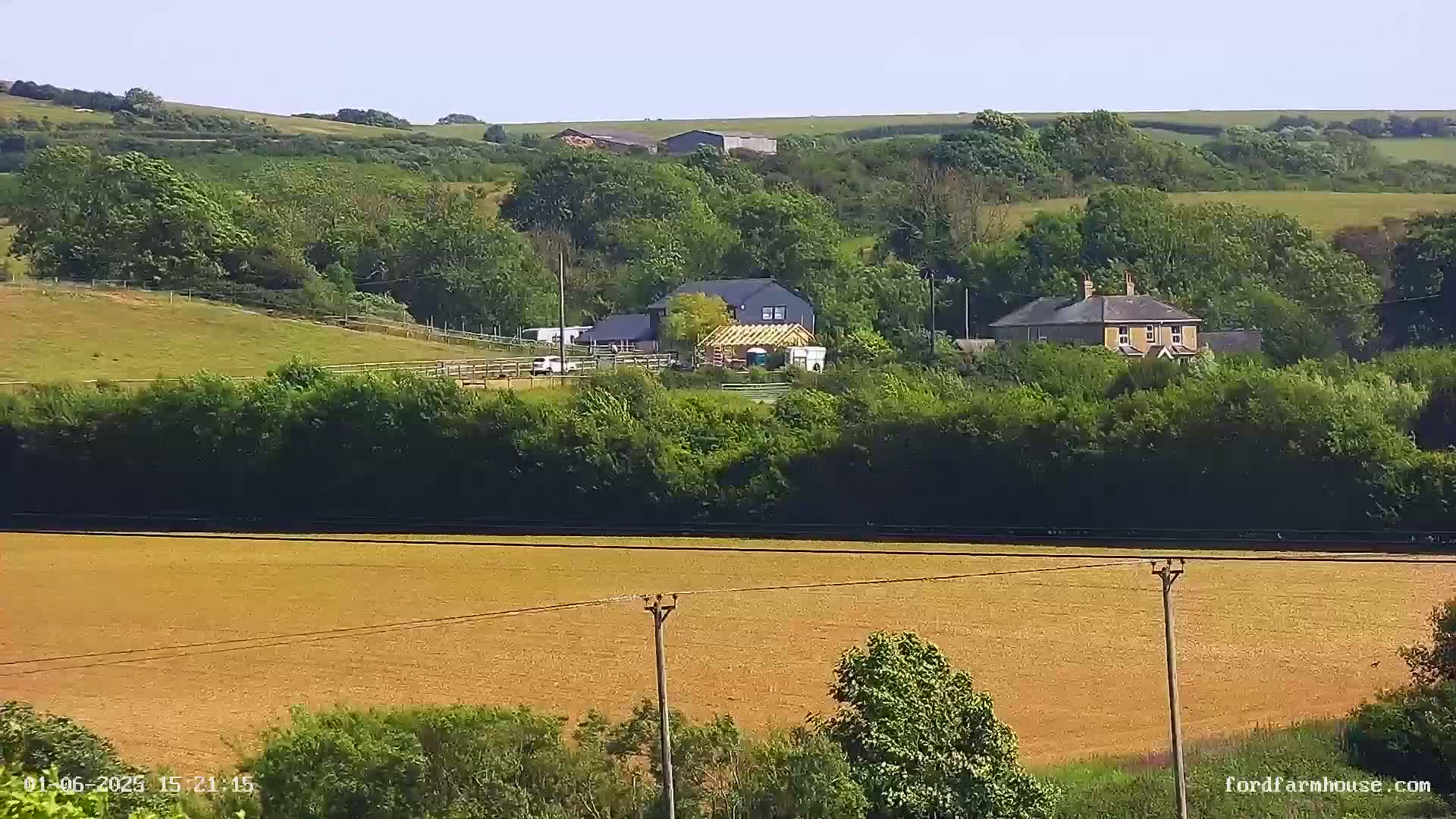 A sprawling rural landscape under sunny skies features a large, tan field in the foreground, a line of trees and shrubs in the midground, and several houses and farm buildings nestled among the hills in the background.