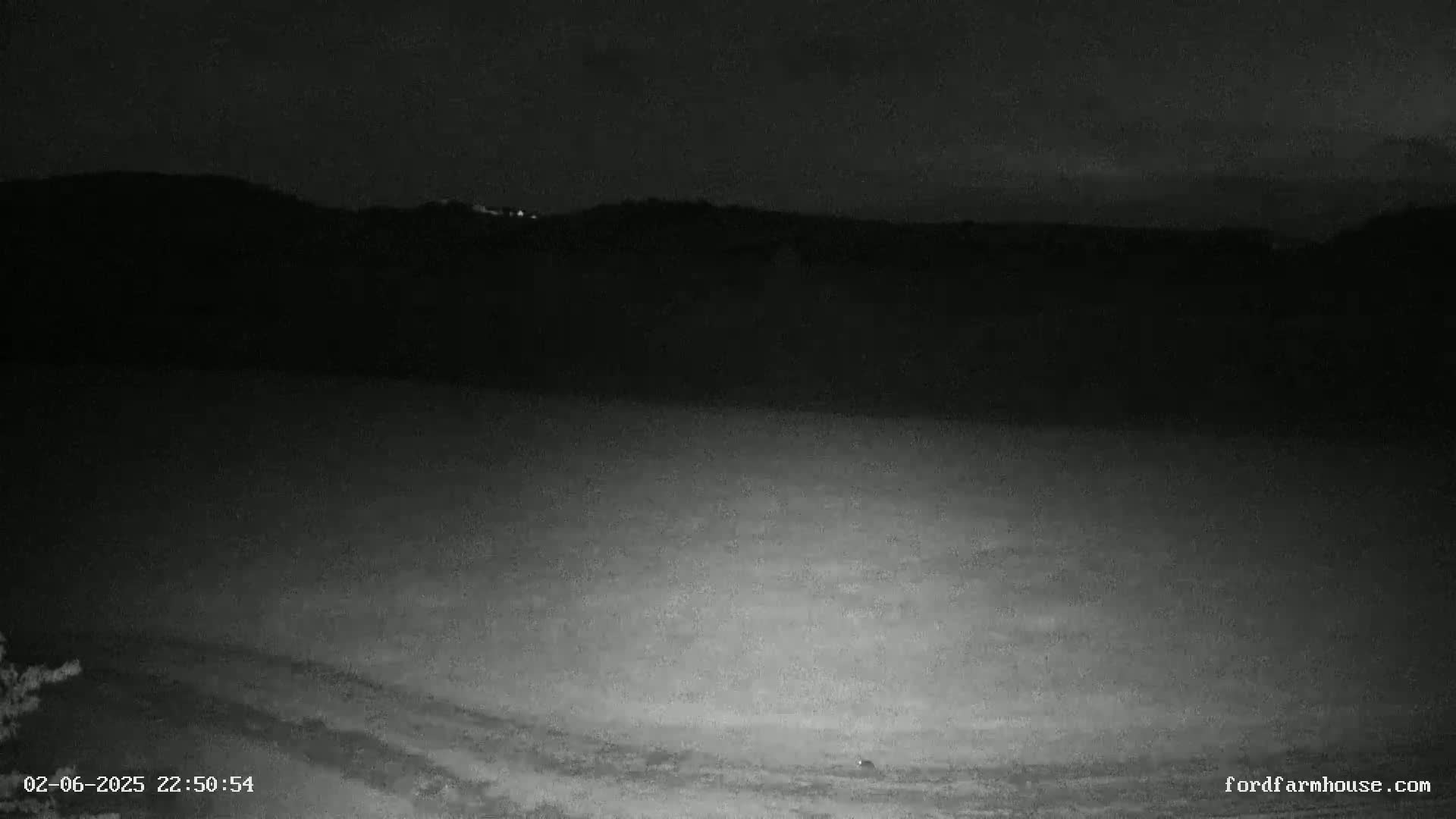 A nighttime, low-light view shows a dark, flat expanse of land leading to a dark hill line under a mostly clear sky.
