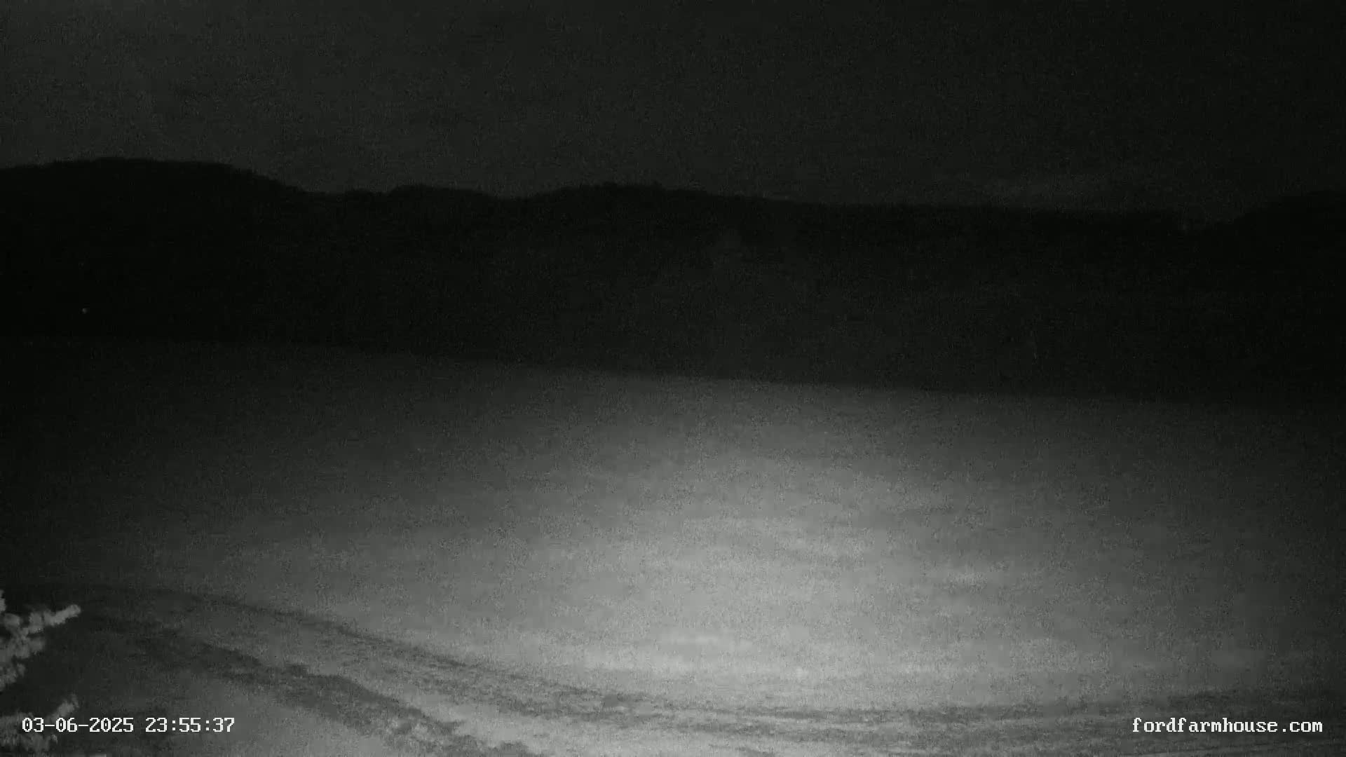 A nighttime, low-light view shows a flat, dark expanse of land extending to a dark silhouette of hills under a mostly clear night sky.