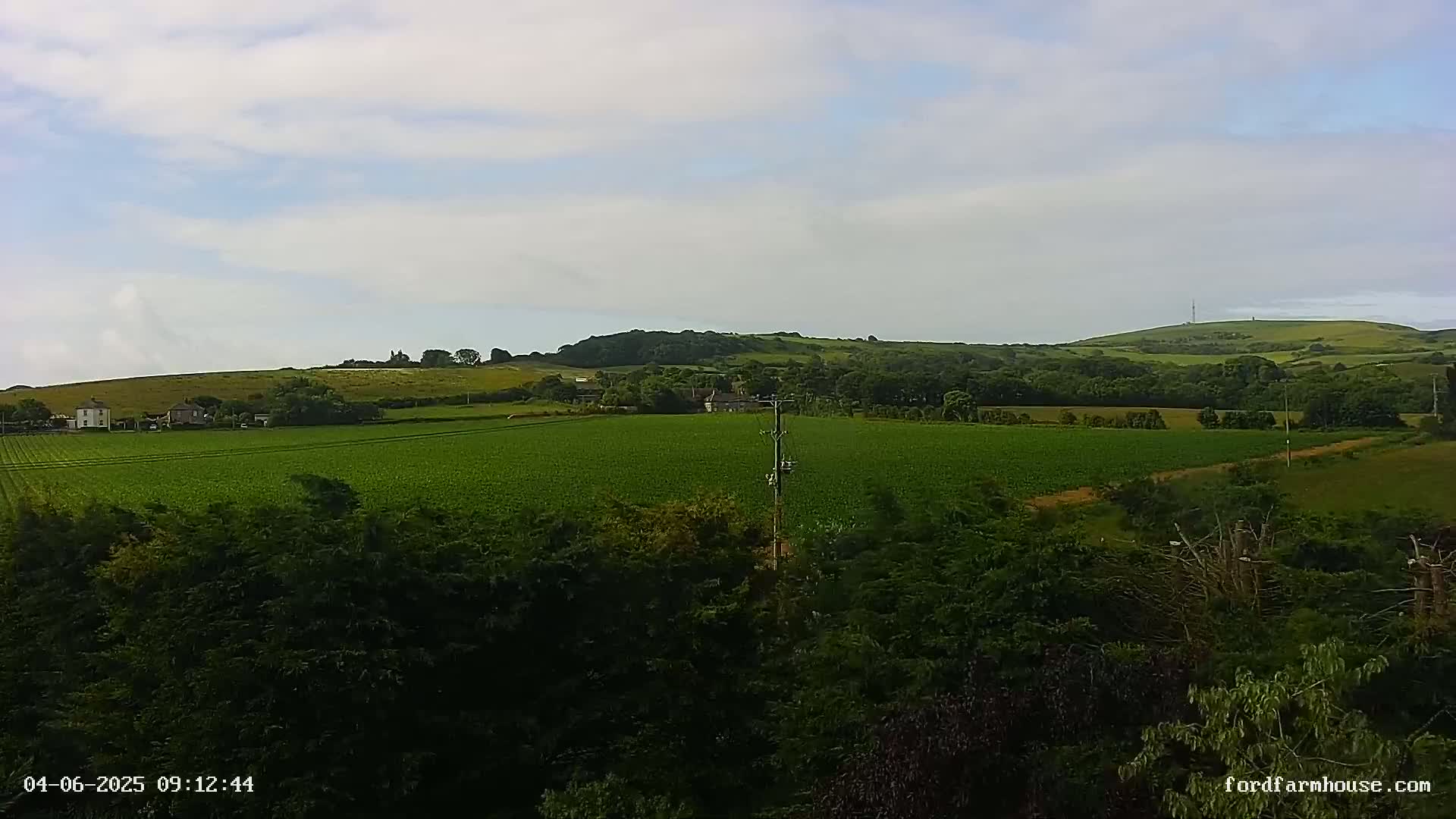 A partly cloudy day reveals a verdant landscape of rolling hills, fields of green crops, and a scattering of houses nestled among the trees.