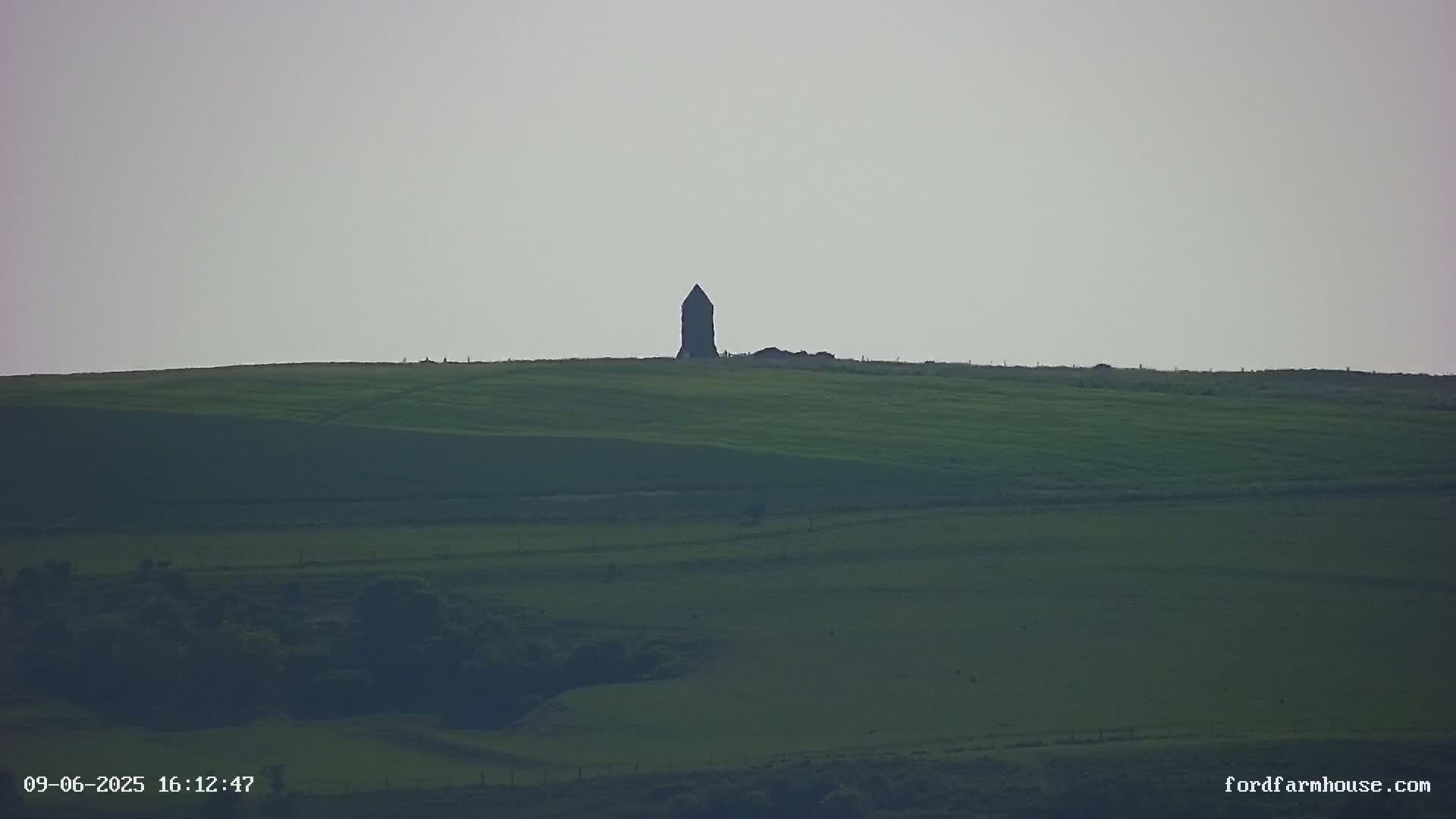 A dark, pointed structure stands atop a gently rolling, green hill under a pale, overcast sky.