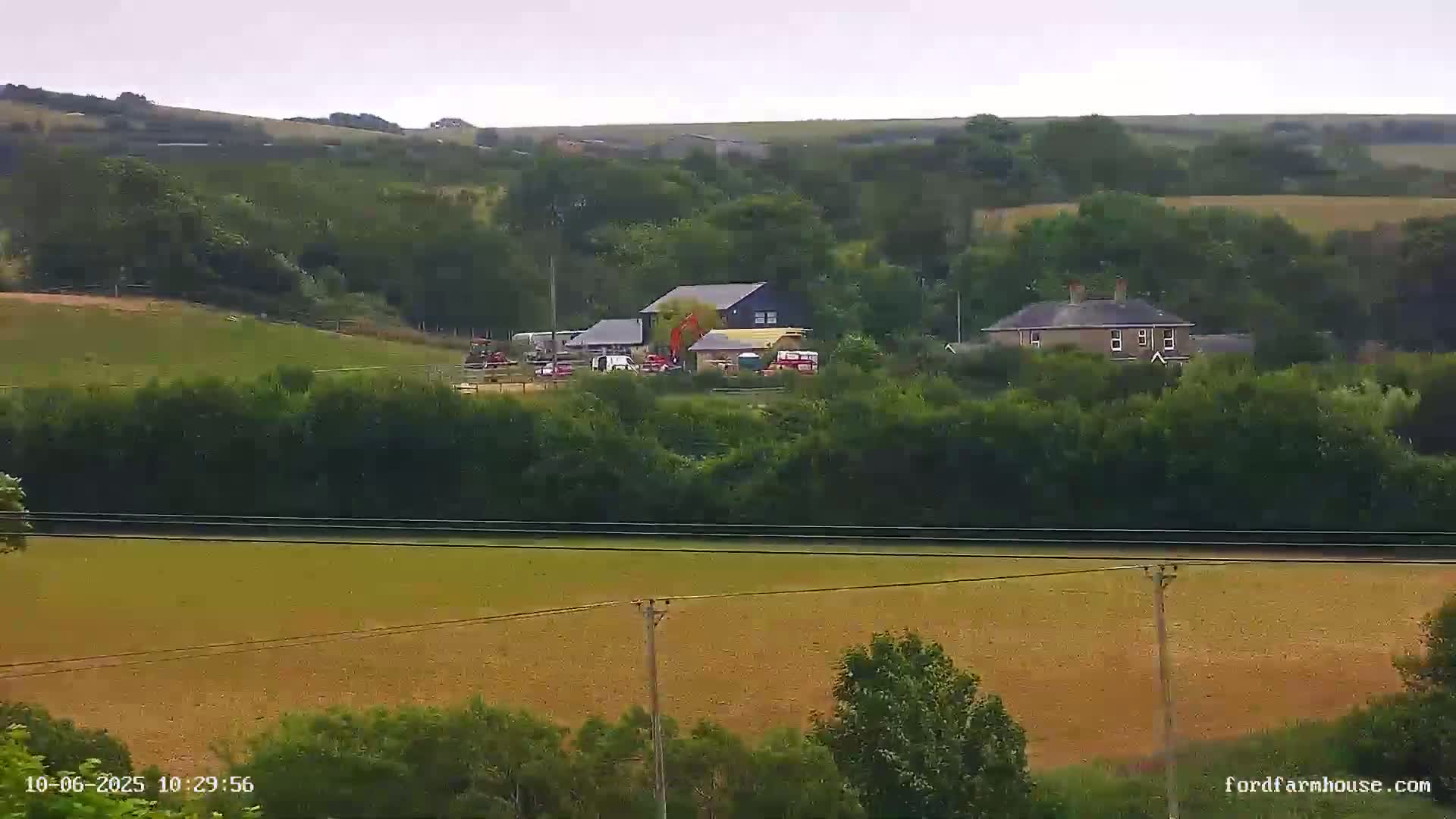 A long shot of a rural landscape under an overcast sky shows several buildings nestled amongst green trees and fields.