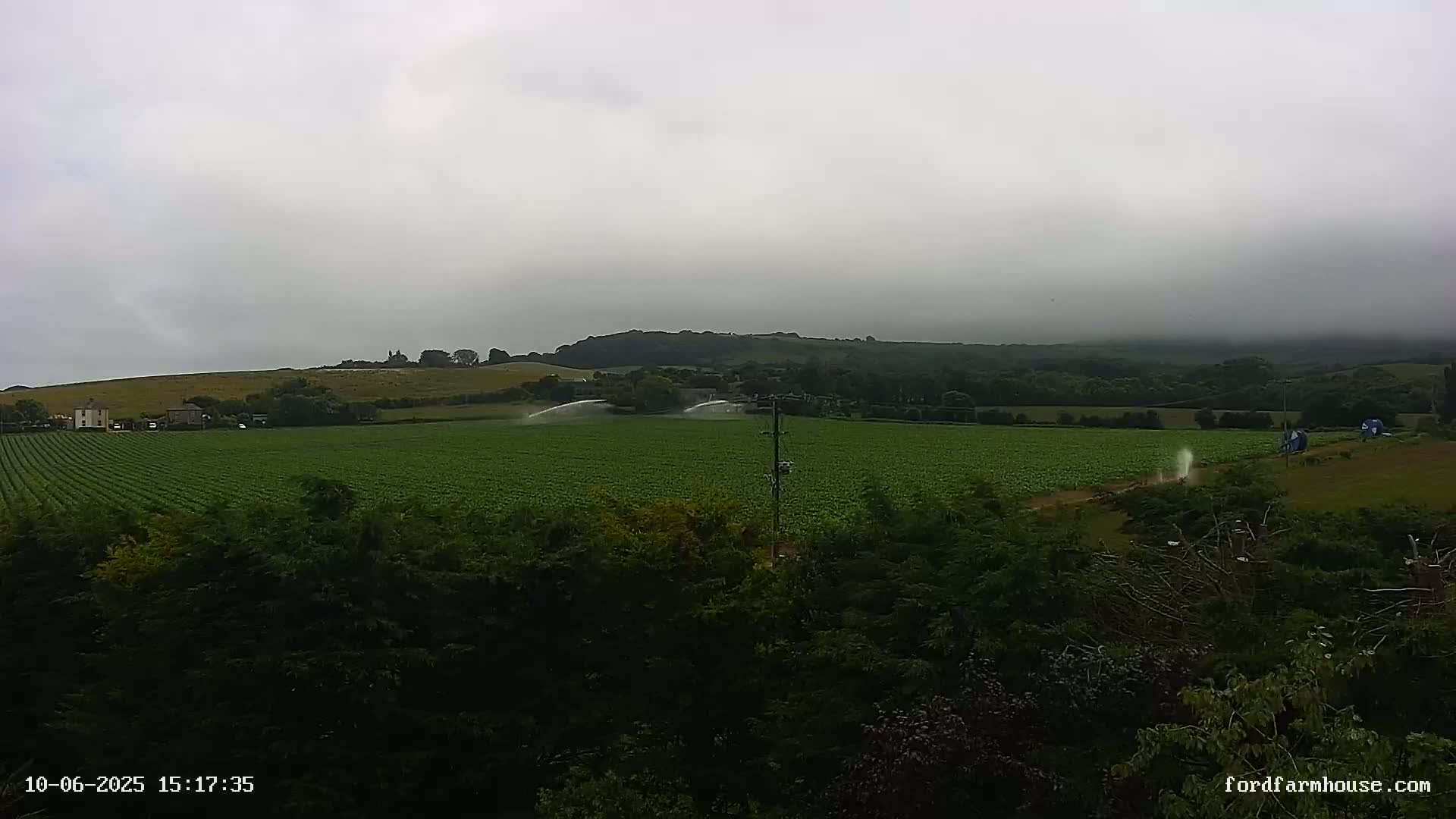 A large, green field is being irrigated under an overcast sky, with a line of trees and hills visible in the background.