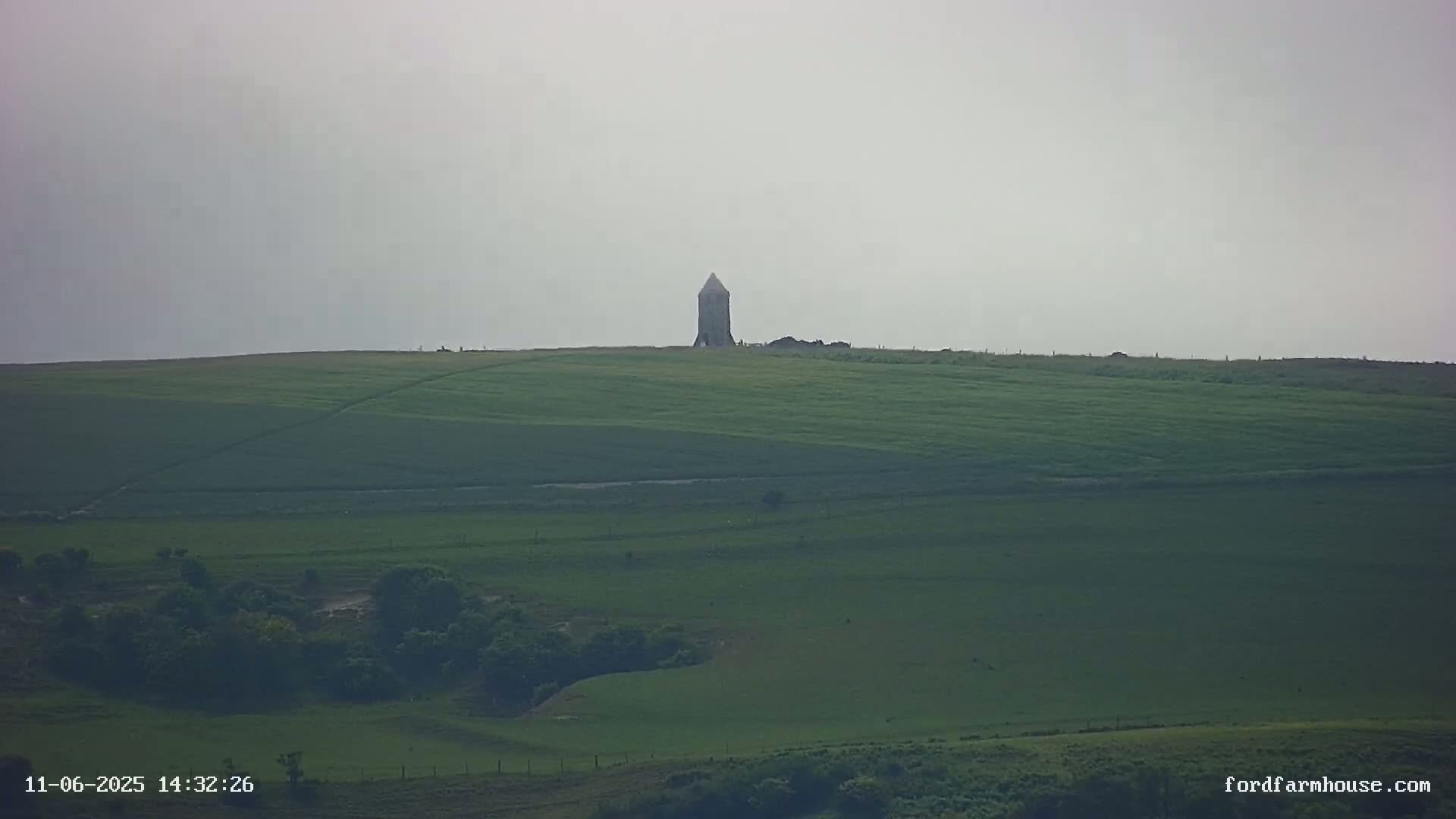 A tall, slender tower stands atop a rolling green hill under an overcast sky.