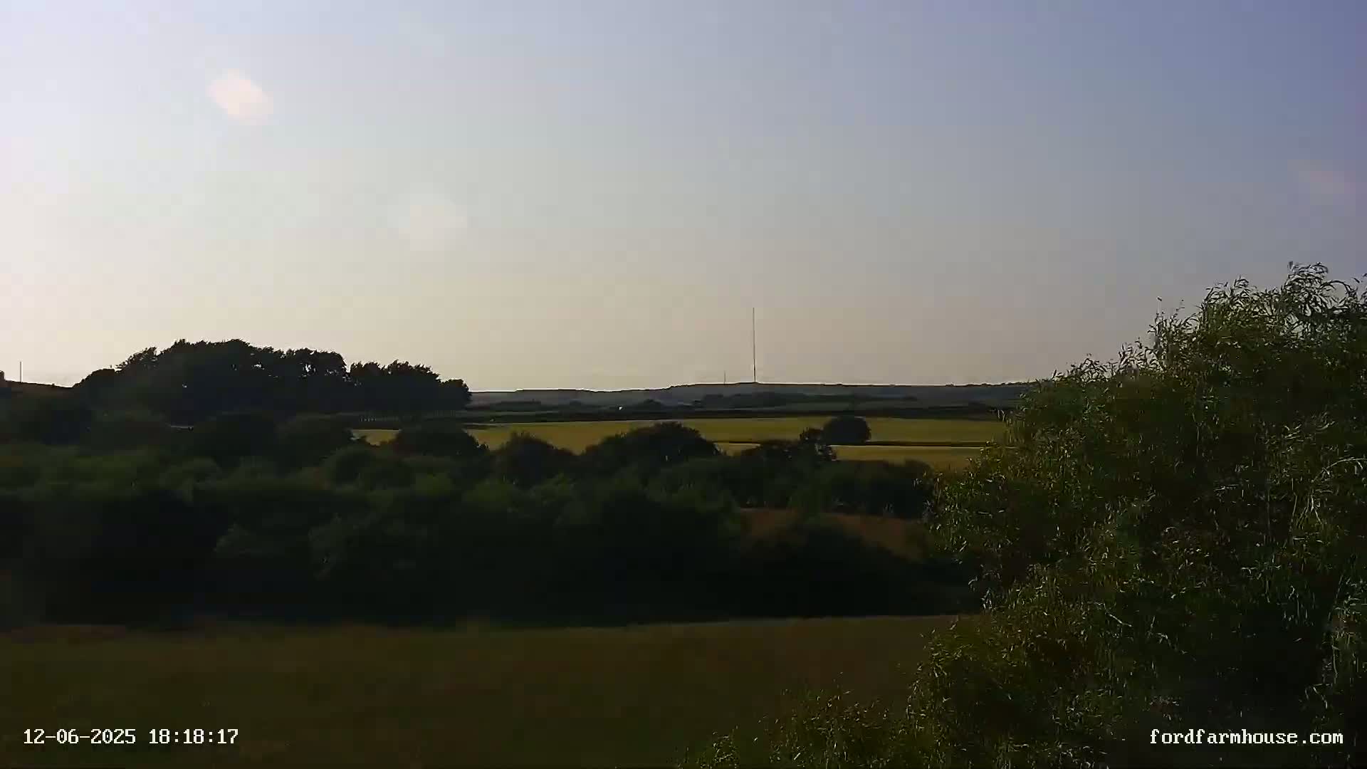 A mostly clear, late afternoon or early evening view of a rural landscape featuring fields, trees, and a distant communications tower.