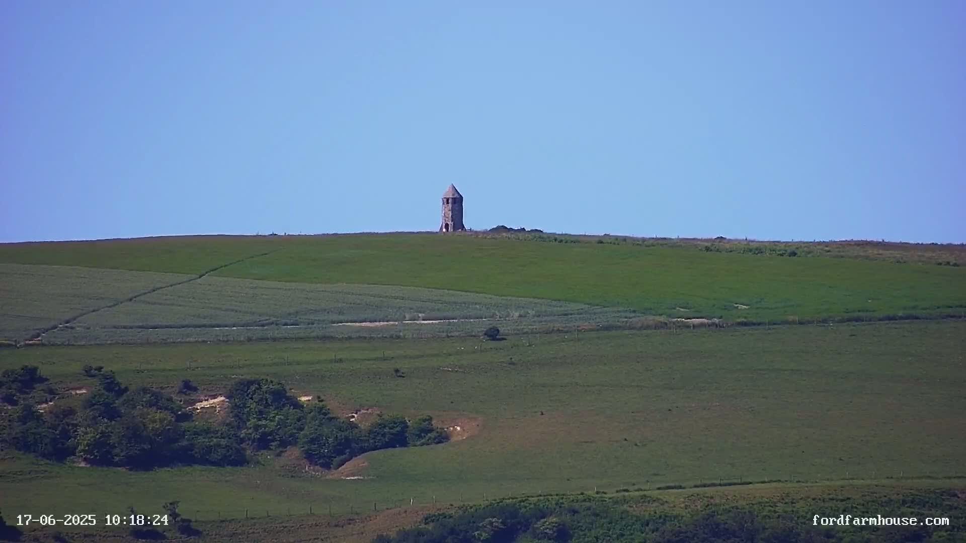 A stone tower stands atop a rolling green hill under a clear blue sky.